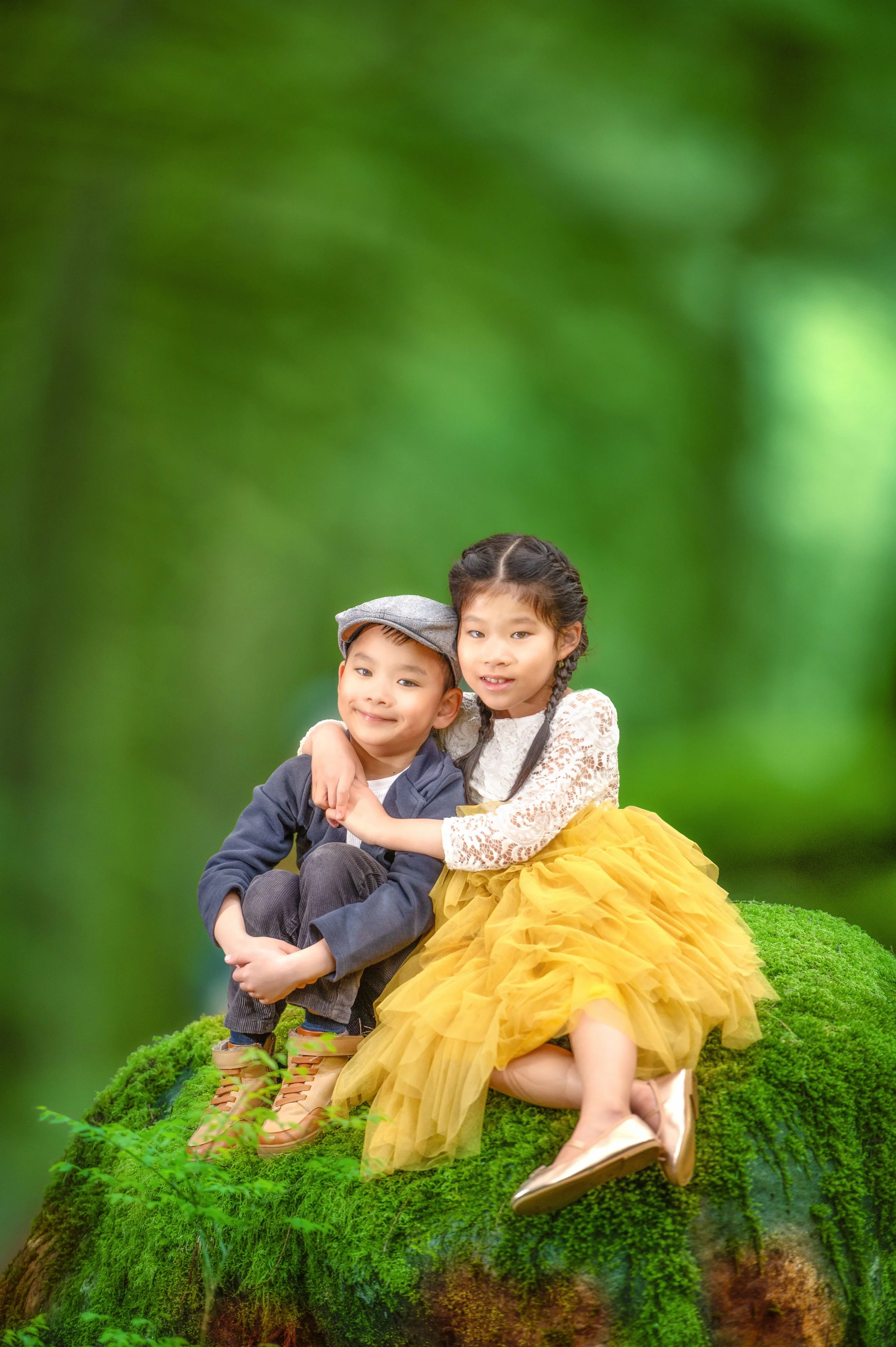 Two children, a boy and a girl, sitting on a moss-covered log in a forest, hugging each other, with a blurred green background.