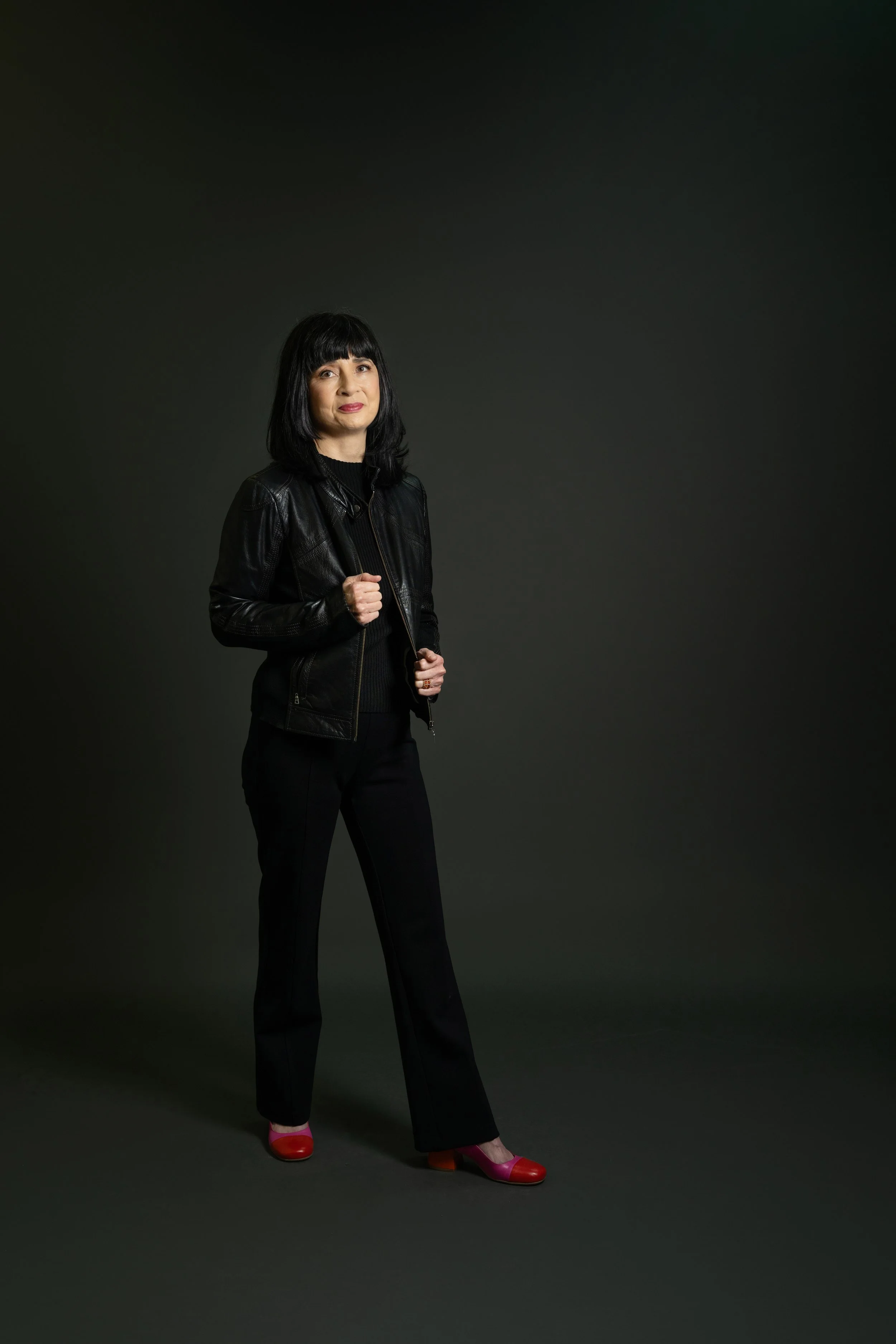 Woman with black bob haircut wearing black leather jacket, black pants, and red high-heeled shoes, standing against a dark gray background.