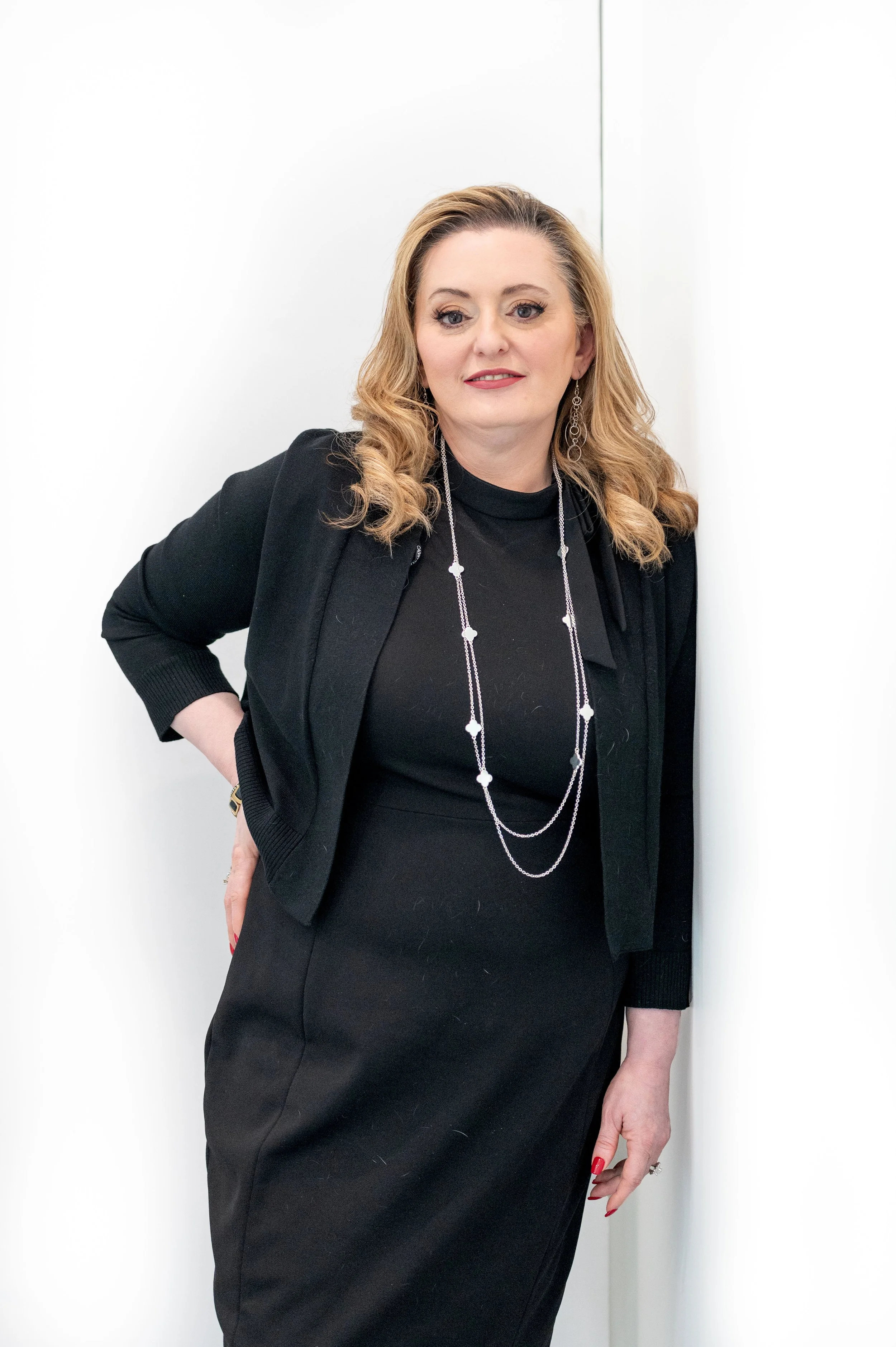 A woman with blonde hair wearing a black dress and black blazer, accessorized with a long silver necklace and earrings, standing against a plain white background.