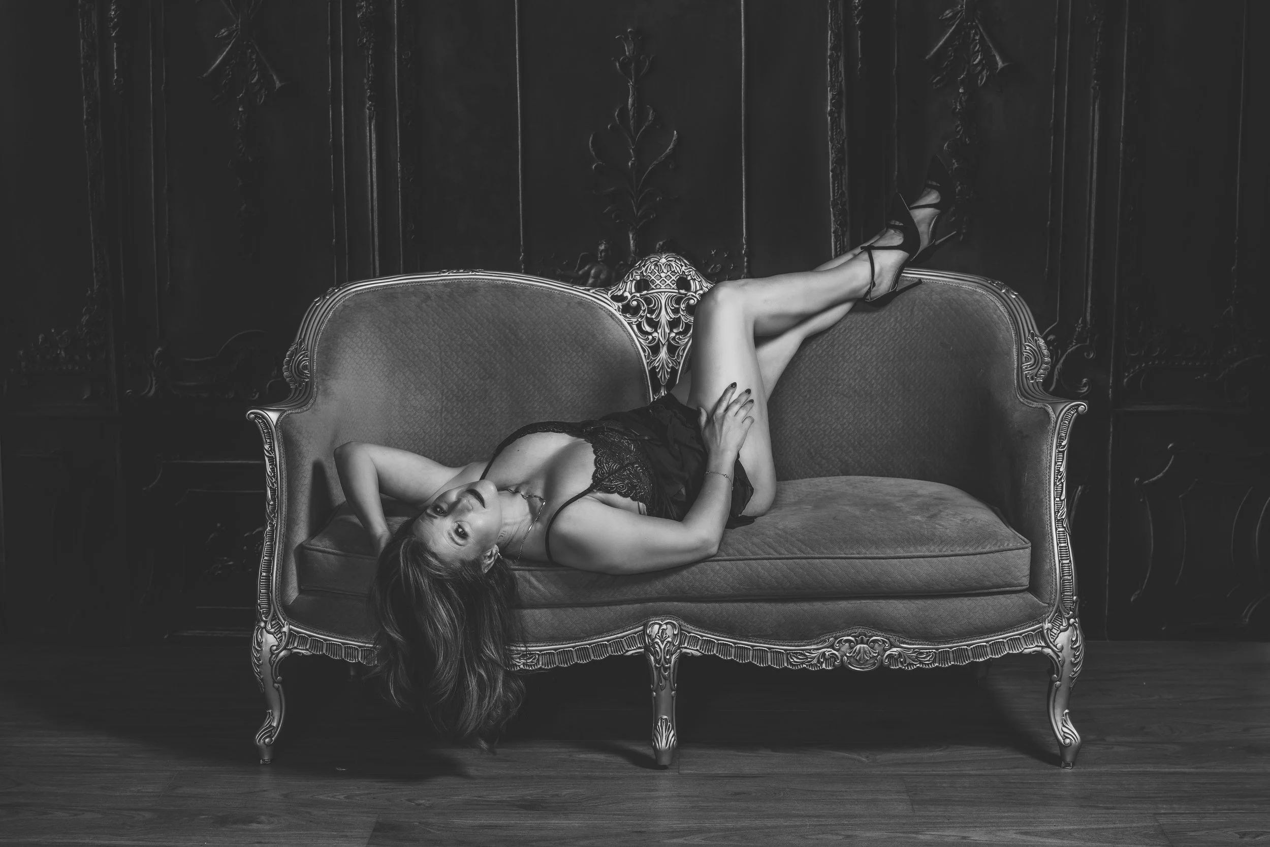 A woman in black lingerie reclining on a vintage sofa in a dark, ornate room.