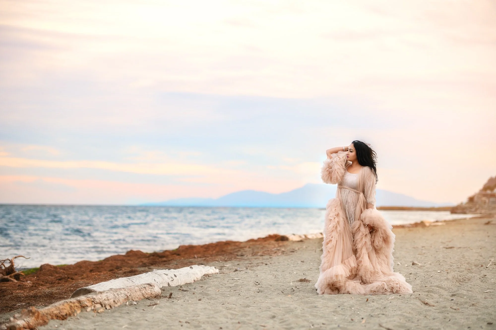 A woman in a flowing beige gown stands on a sandy beach near the ocean, with a pastel-colored sky and mountains in the background.