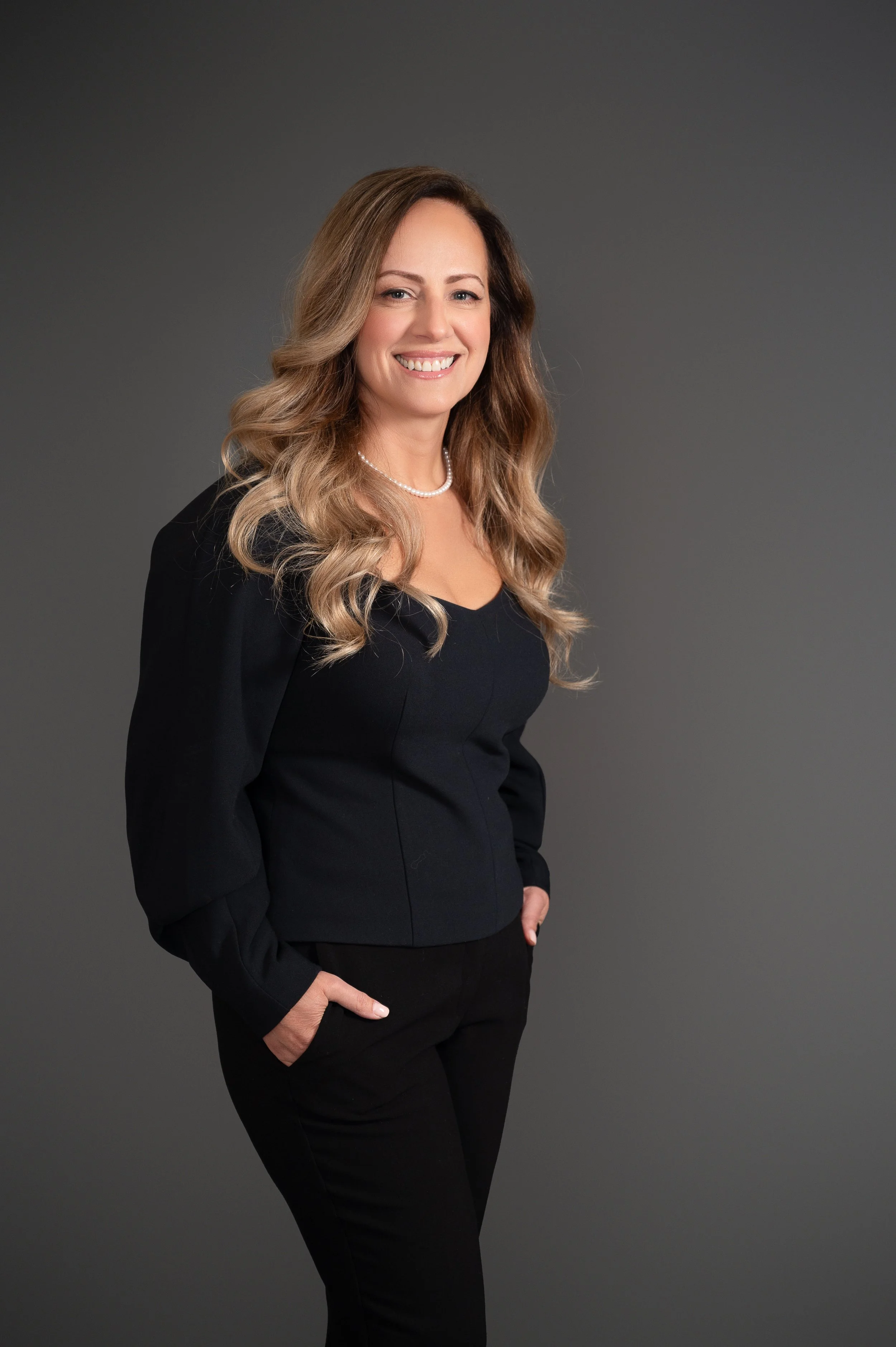 Professional woman with wavy blonde hair wearing a black blazer, black pants, and a pearl necklace, smiling against a gray background.