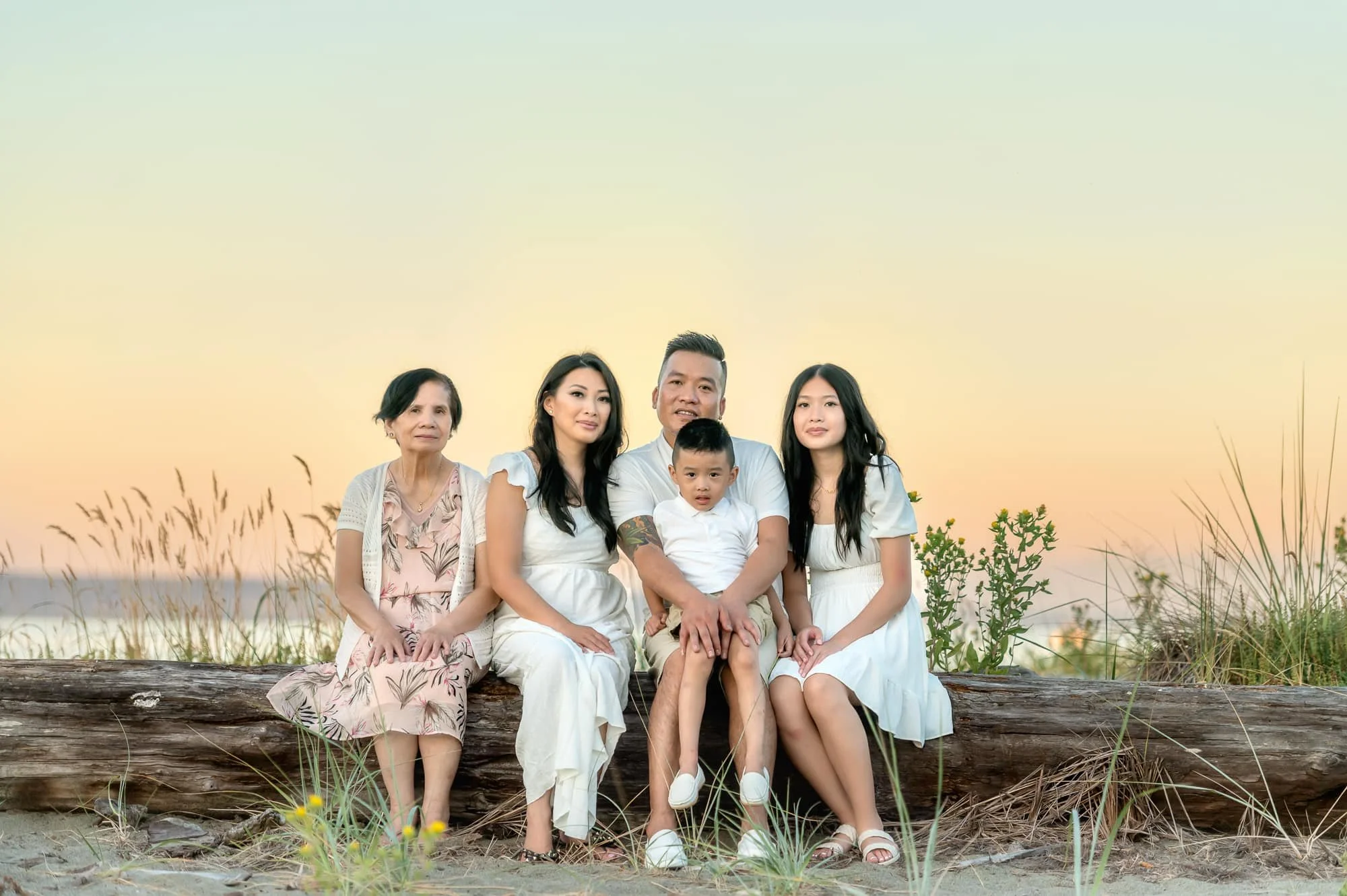 A multi-generational Asian family of five sitting on a log at the beach during sunset, with tall grass and the ocean in the background.