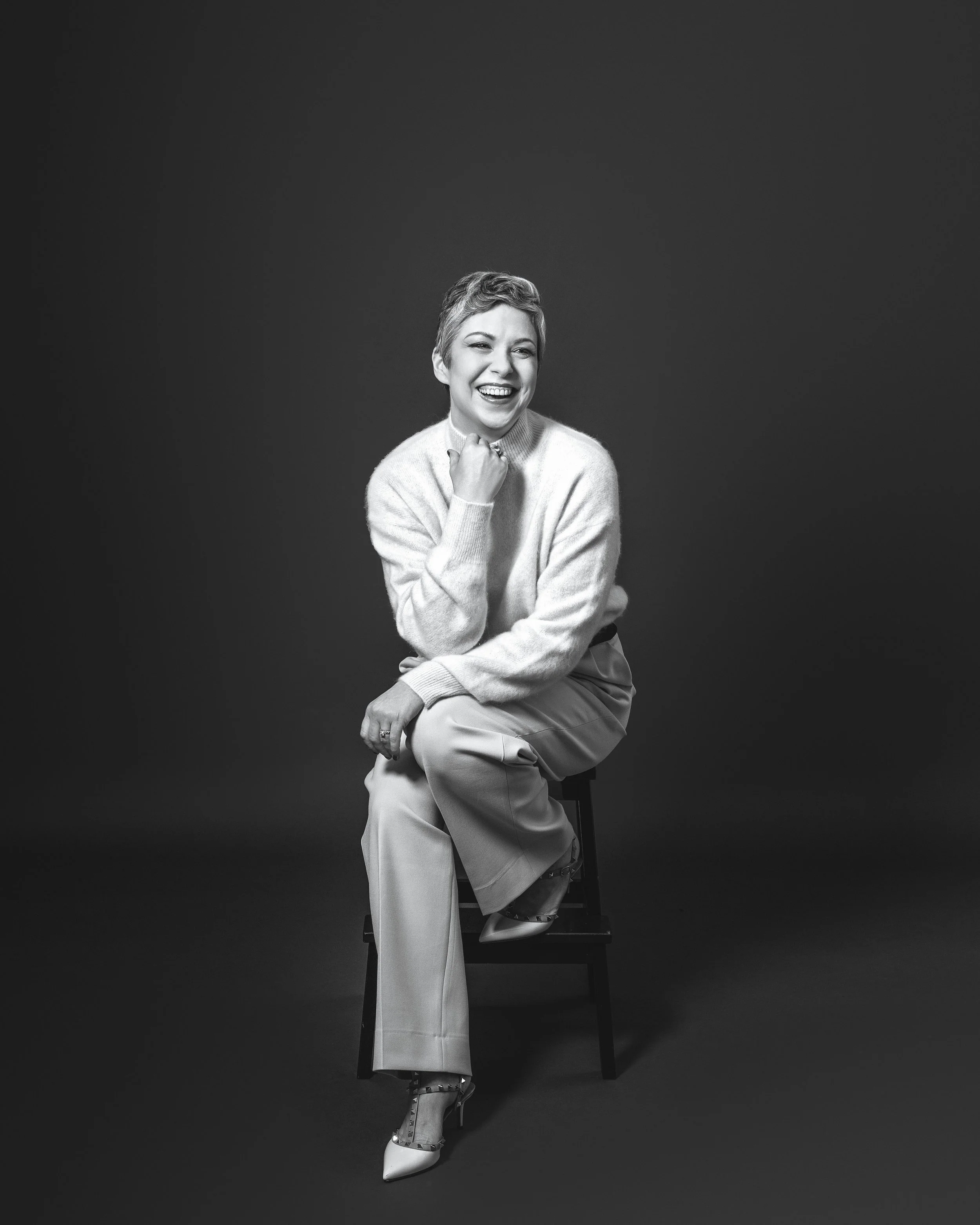 Black and white portrait of a woman sitting on a chair, smiling and laughing with her hand near her chin, wearing a cozy sweater and wide-leg pants, against a plain dark background.
