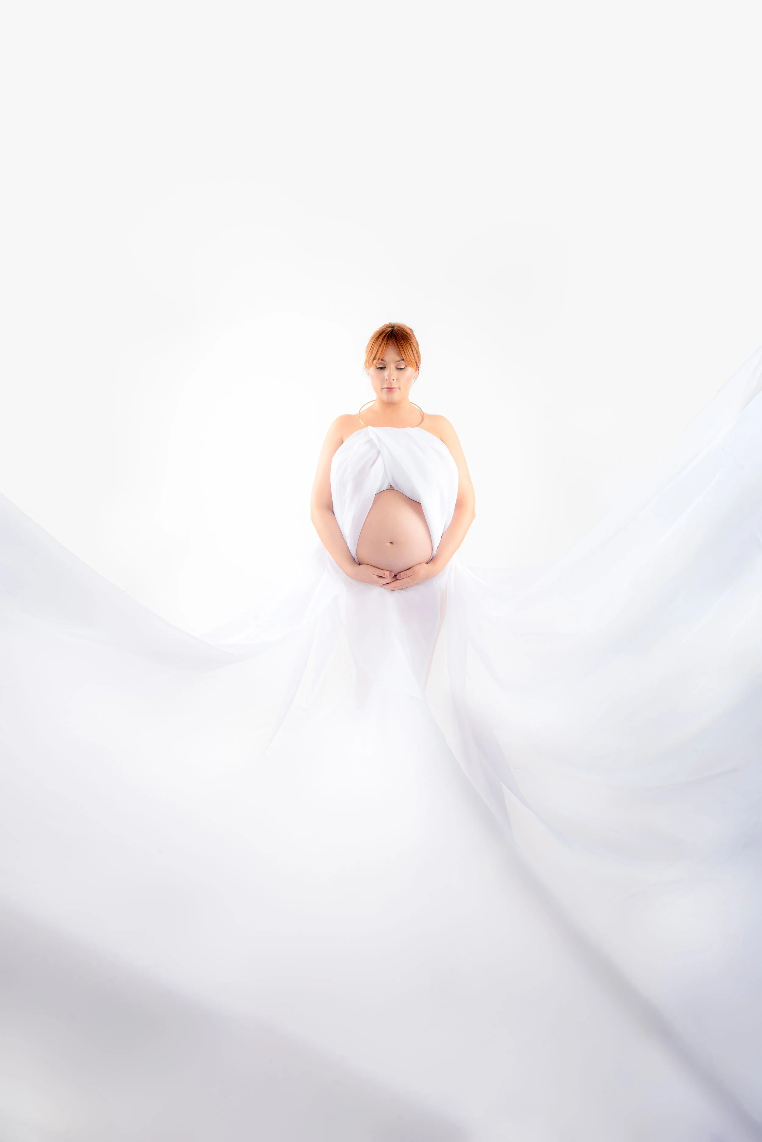 Pregnant woman in white lying on a white fabric background, with her hands on her belly.