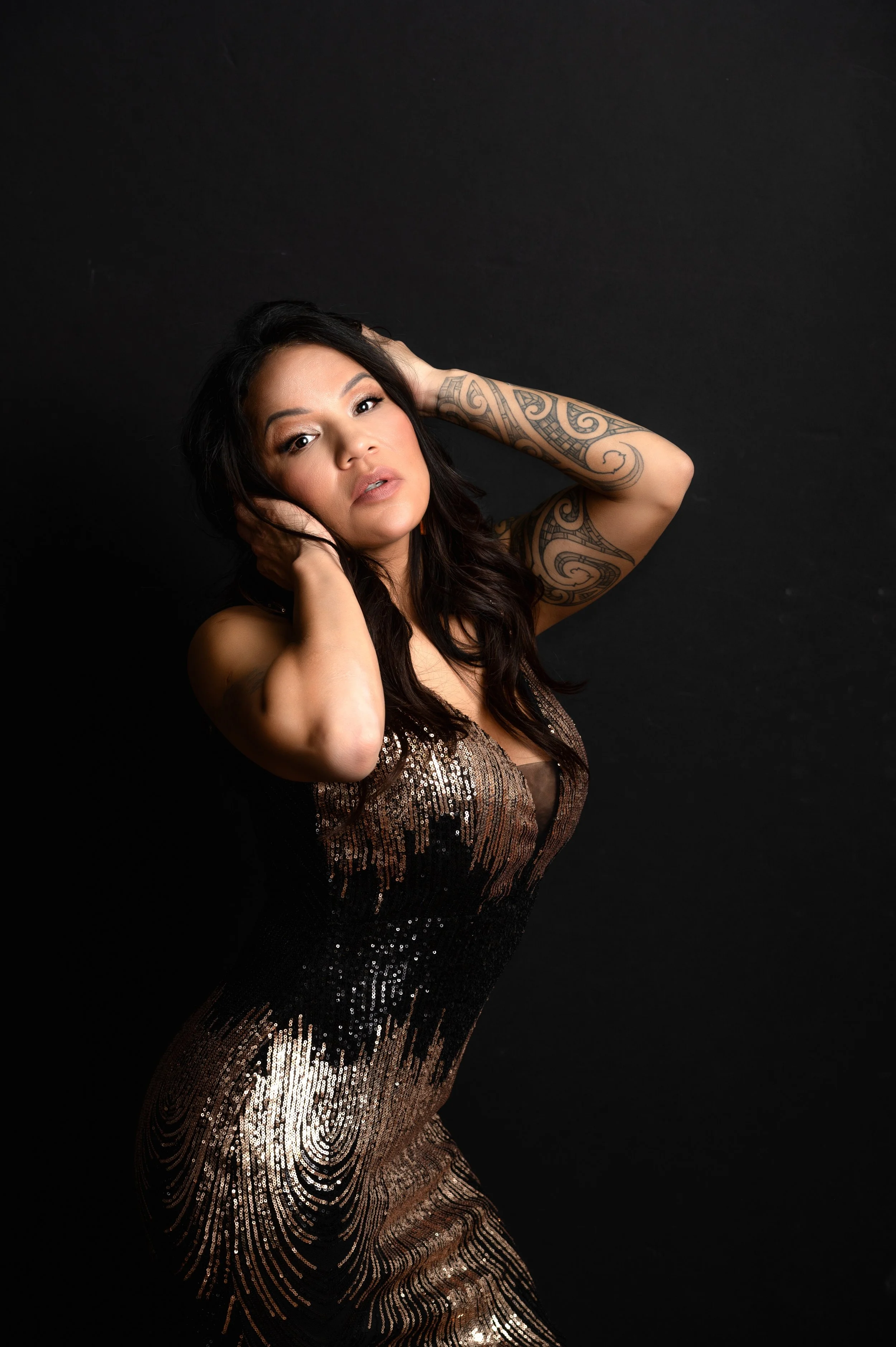 A woman with long dark hair, wearing a sparkly, patterned dress, poses with her hands near her head against a black background.