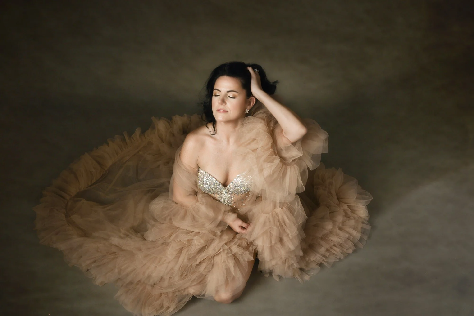 Woman wearing a beige tulle gown with a bejeweled bodice, sitting with eyes closed and her hand in her hair against a neutral background.