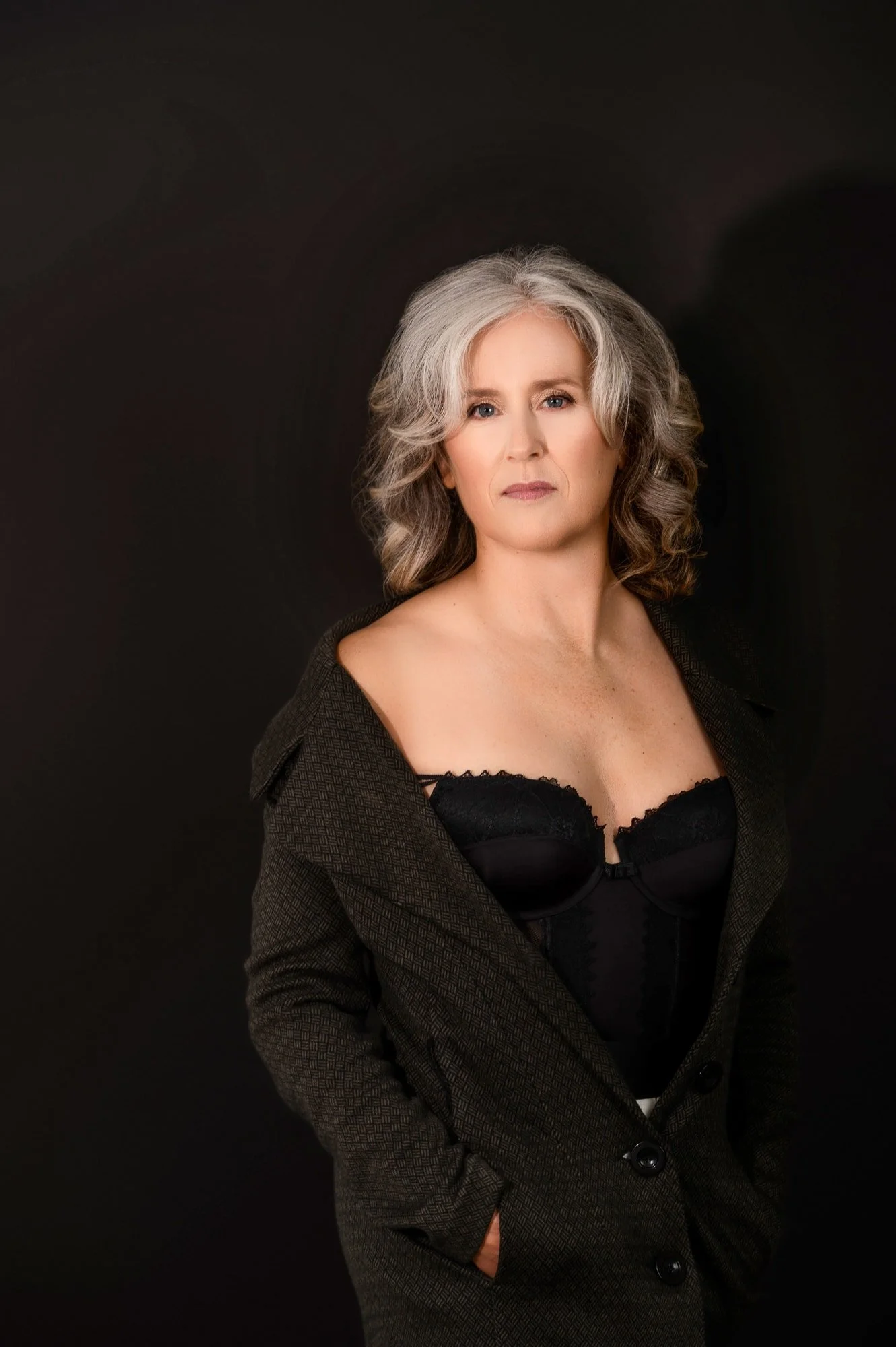 Portrait of a mature woman with gray, curly hair wearing a dark blazer and a black lace bustier against a black background.