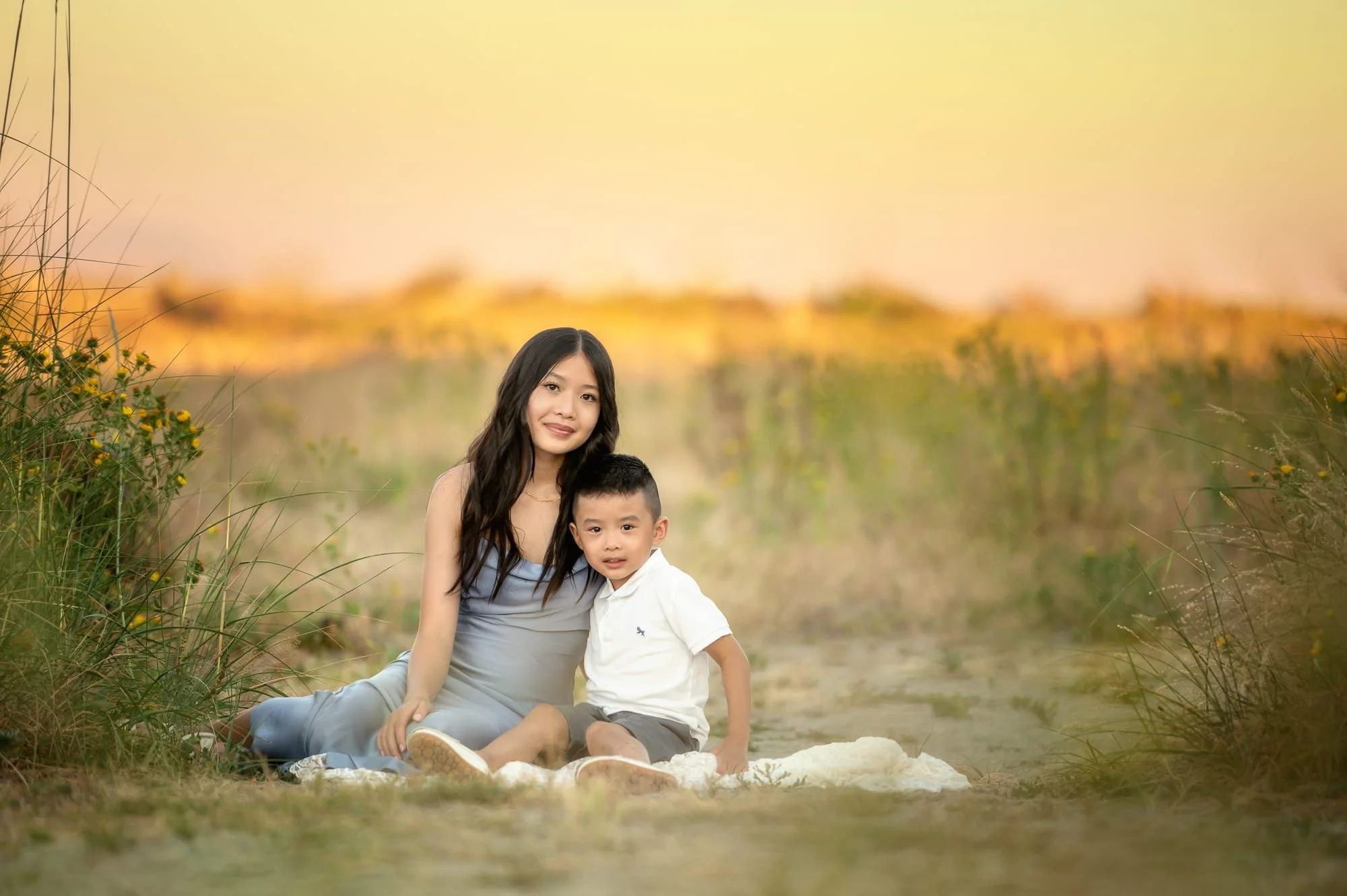A woman and a young boy sitting on a blanket outdoors in a natural setting with tall grass and yellow flowers, during sunset or golden hour, with a blurred landscape in the background.