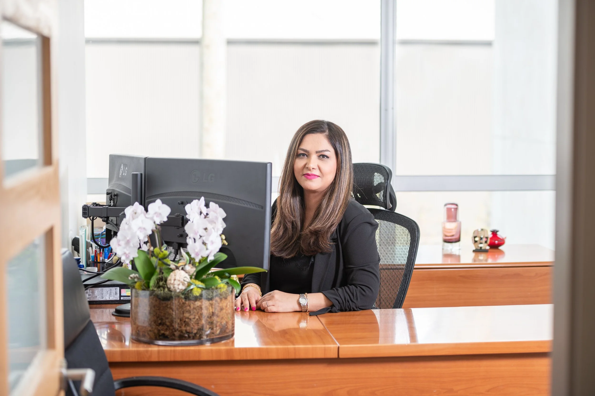 A woman with long brown hair wearing a black blazer sitting at a wooden desk in an office, with a computer monitor in front of her and decorative items in the background.