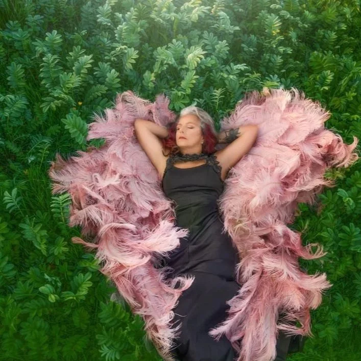 Woman lying on her back on green grass, surrounded by a heart-shaped arrangement of pink feathers, wearing a black dress and resting with eyes closed.