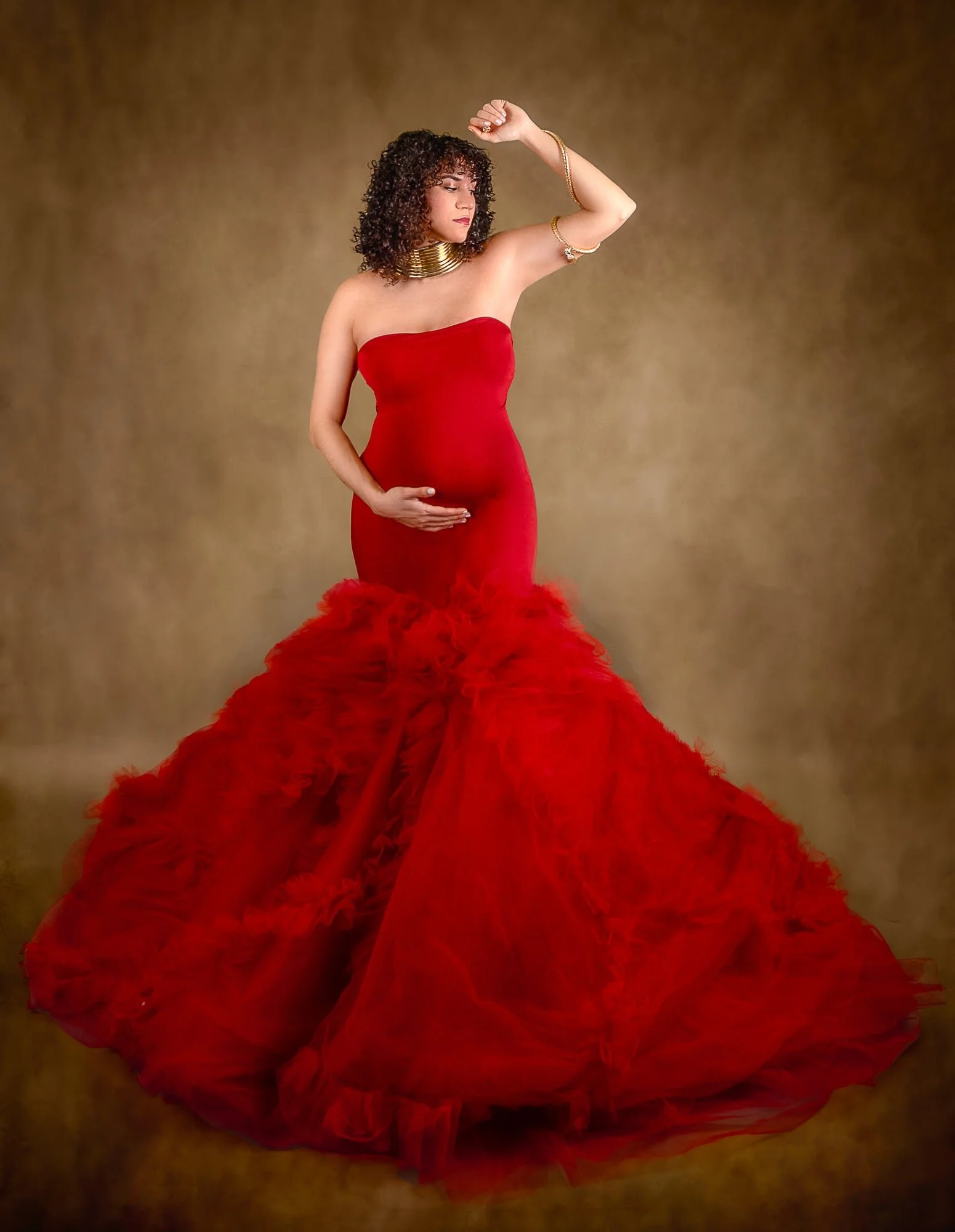 A pregnant woman in a strapless red wedding gown with a ruffled skirt, wearing gold jewelry, stands against a brown textured background.