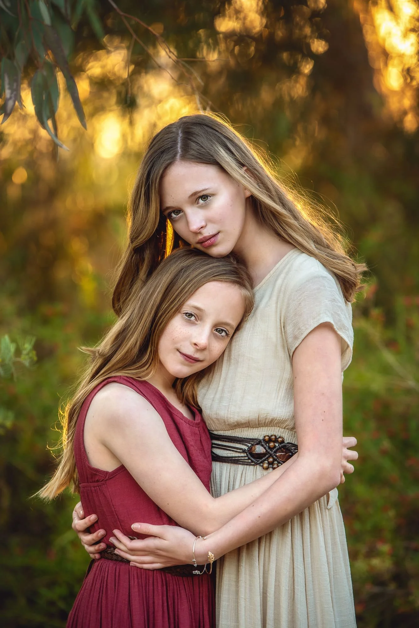 Two young girls standing close together outdoors during sunset, with one wearing a beige dress and the other wearing a red dress, both with long hair and gentle expressions.