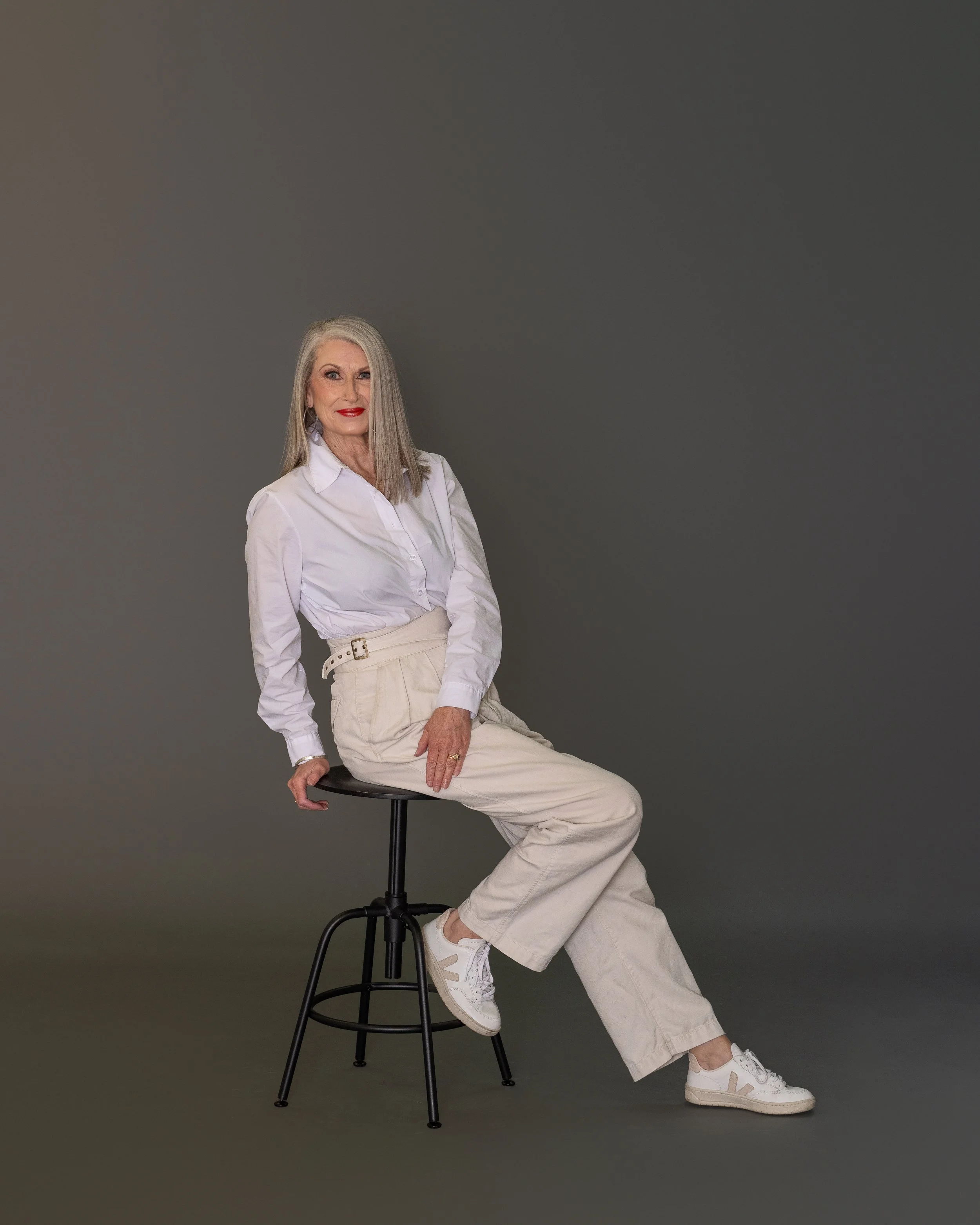 A woman with long gray hair wearing a white button-up shirt and beige wide-leg pants, sitting on a black stool against a plain gray background.