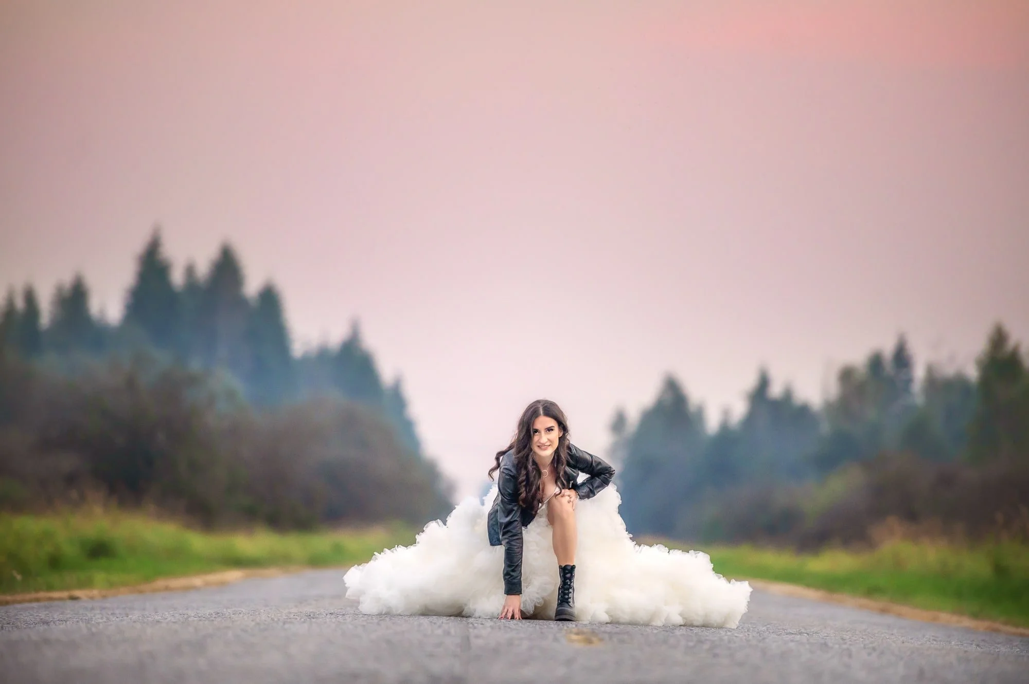 A woman with long dark hair wearing a black leather jacket, white dress, and black boots kneels on an empty road, smiling at the camera with a forest and pinkish sky in the background.