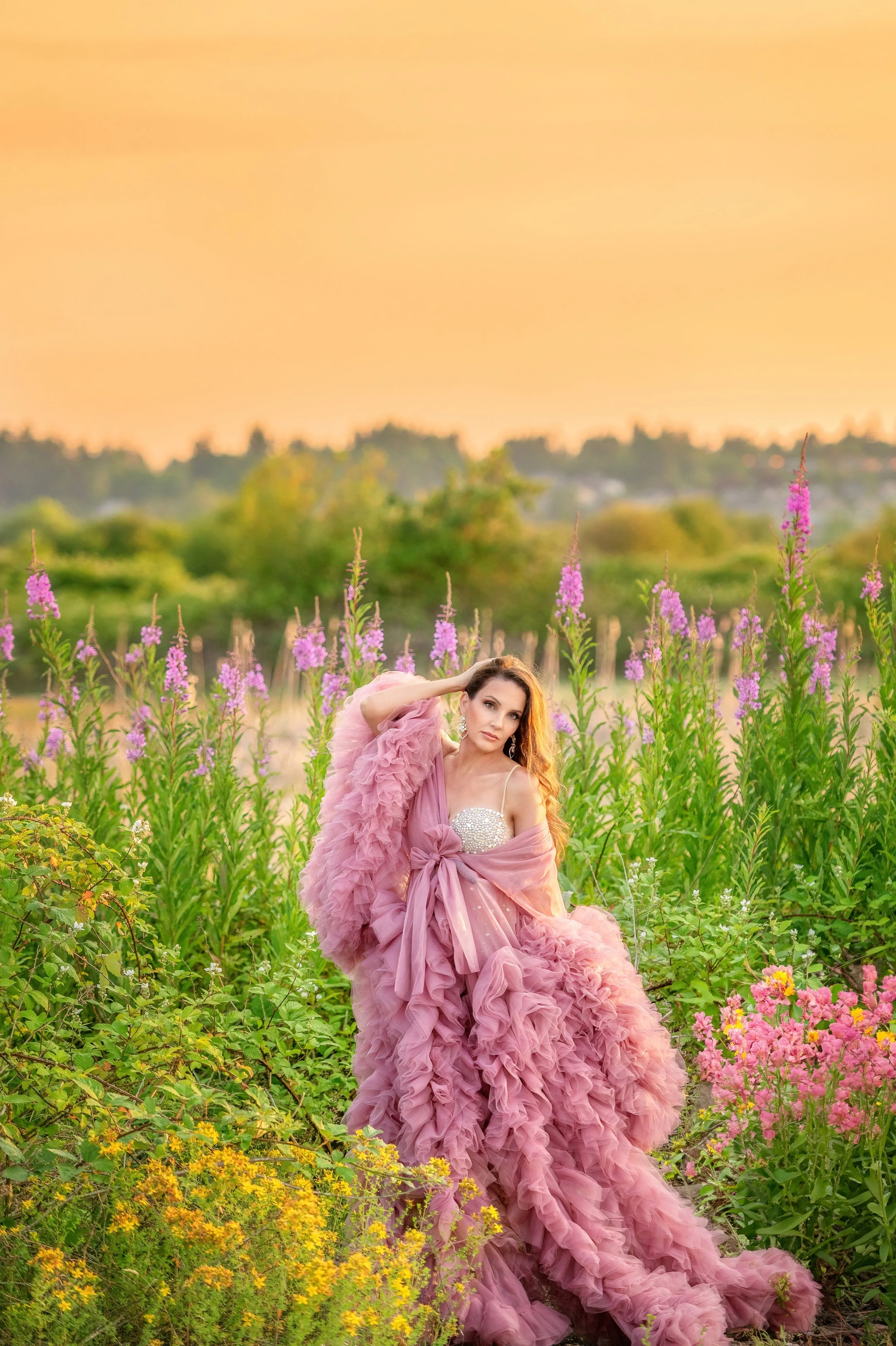 A woman in a pink ruffled gown standing in a field of purple and pink flowers during sunset.