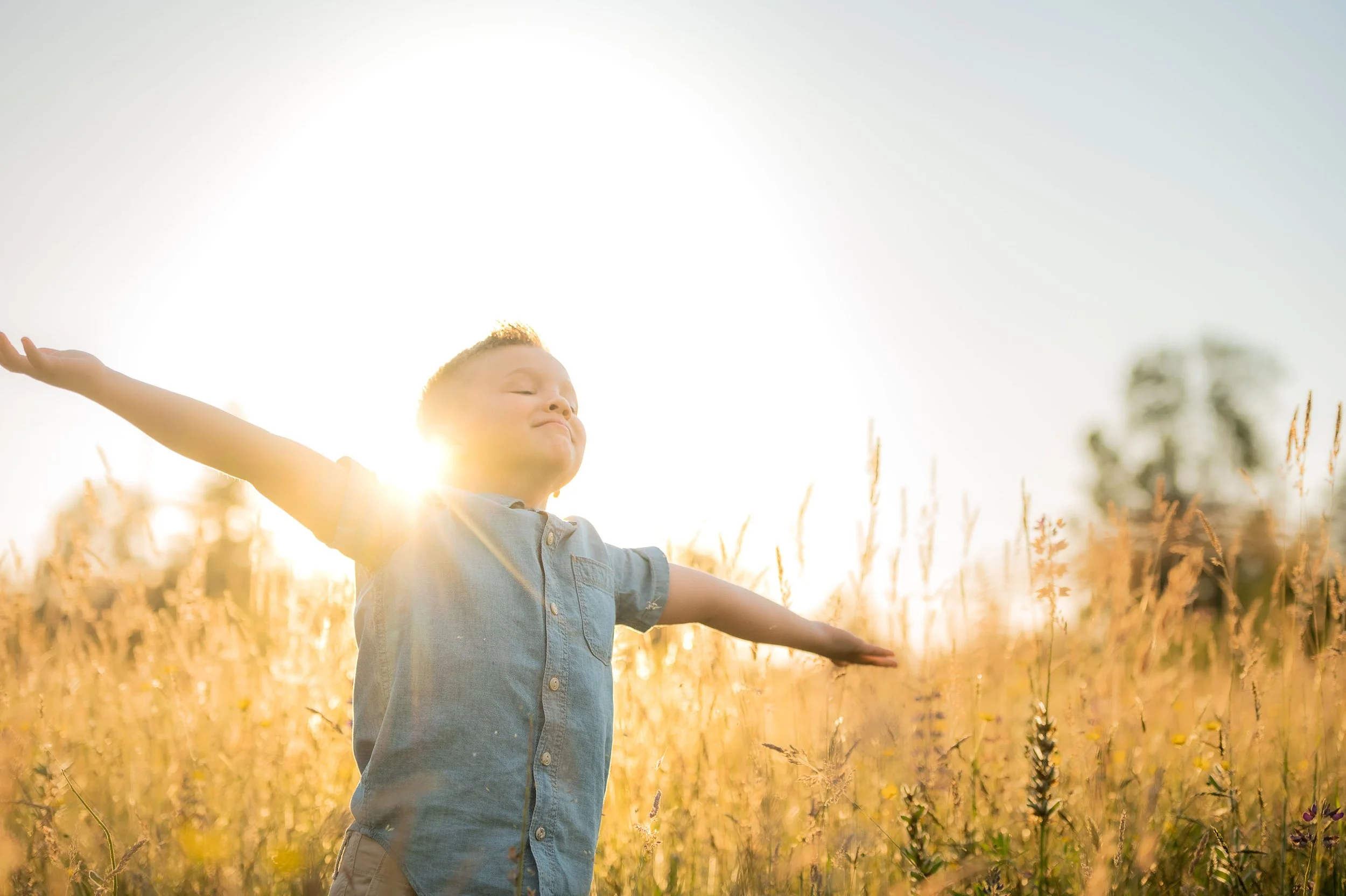 A young boy standing in a field of tall grass with arms outstretched, basking in the sunlight during a clear day.