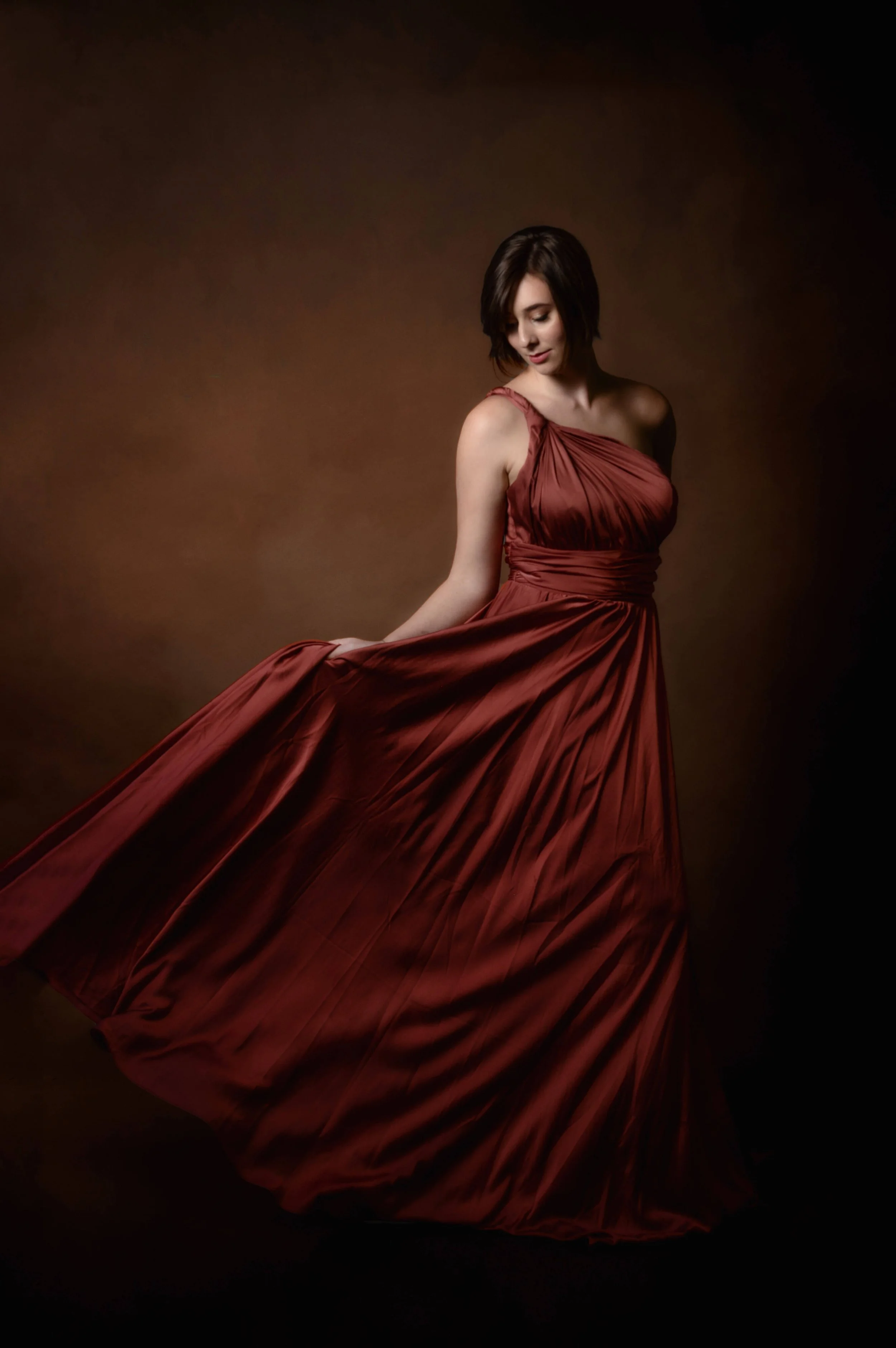 A woman in a long red gown standing against a dark background, holding the hem of her dress and looking down.