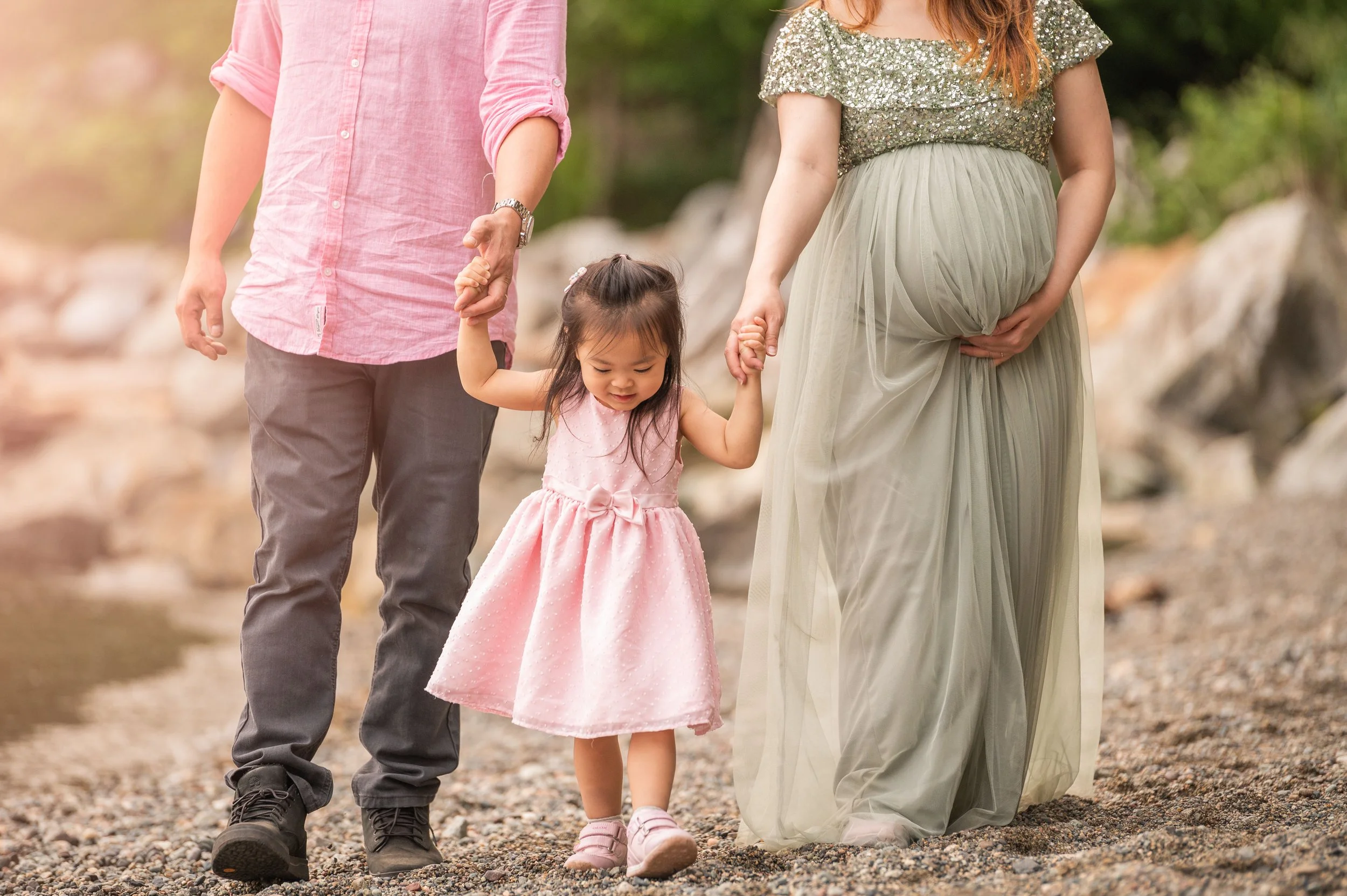 A family walking hand in hand along a rocky beach. The child, wearing a pink dress, is being held by her mother in a floral top and flowing pants, while the father in a pink shirt and gray pants holds her other hand.