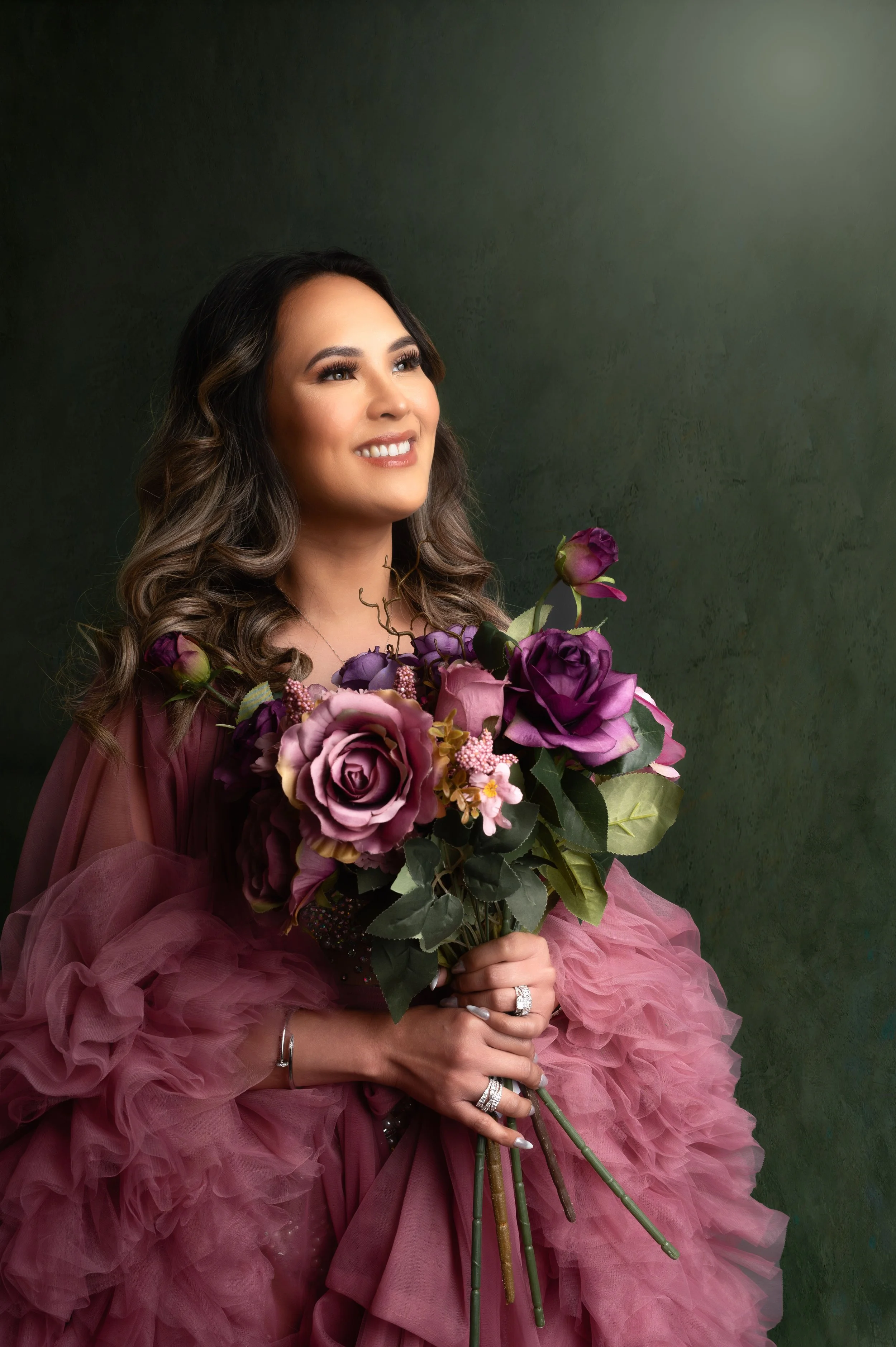 A woman with long wavy hair smiling, holding a bouquet of purple and pink flowers, wearing a pink ruffled gown, against a dark textured background.