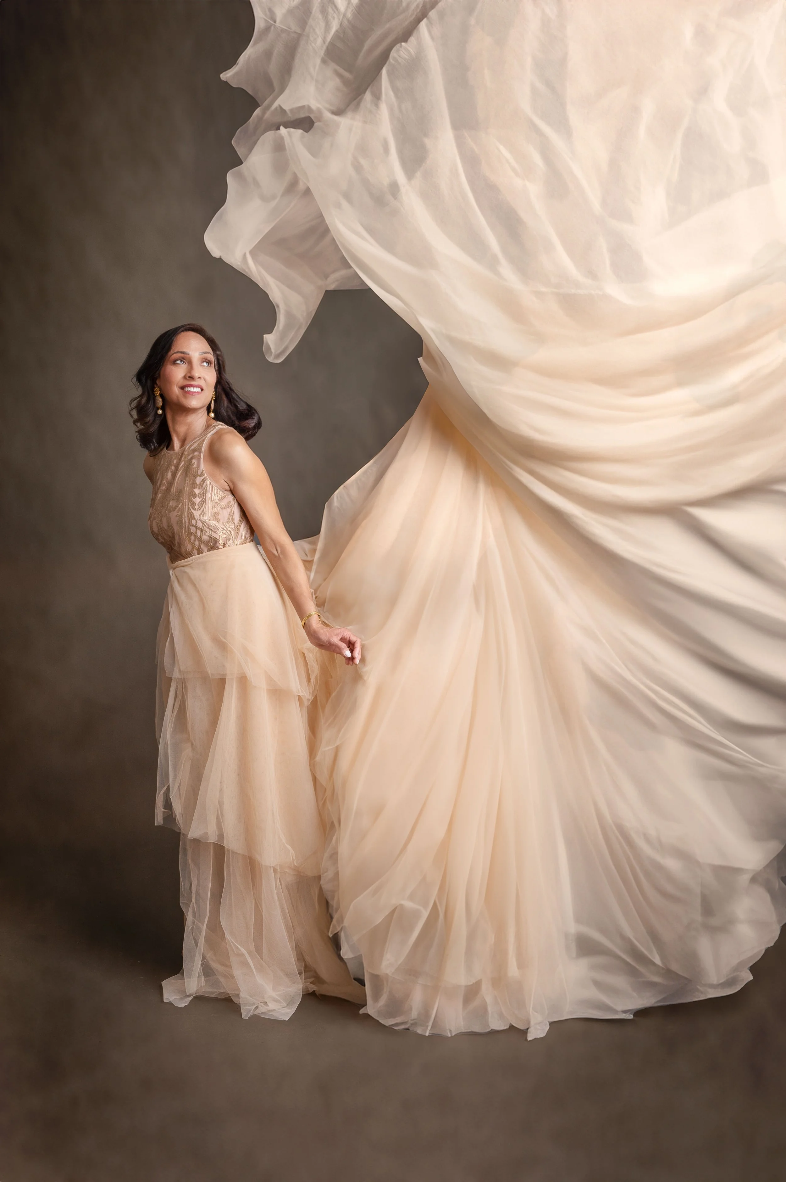 A woman in a beige dress holding the train of a voluminous beige gown with ruffles and layers, against a gray background.