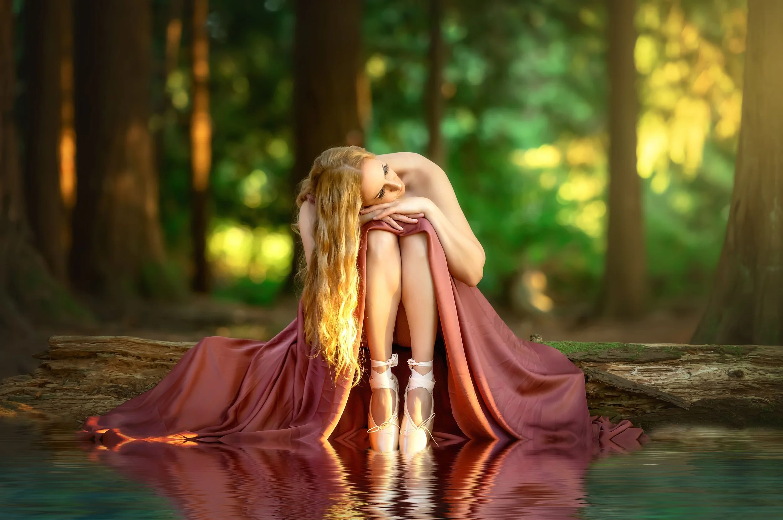 A woman with long blonde hair, wearing ballet slippers, sitting on a log by a calm body of water in a forest, covered partially with a flowing reddish-brown fabric, in a contemplative pose.