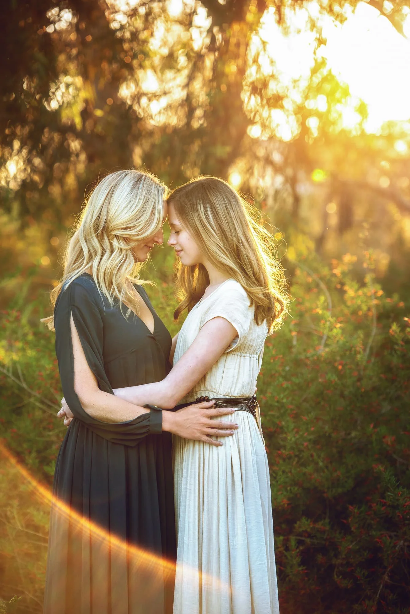 Two women standing close together outdoors during sunset, their foreheads touching. The woman on the left has blonde hair and is wearing a black dress, while the woman on the right has light brown hair and is wearing a light-colored dress. They are e