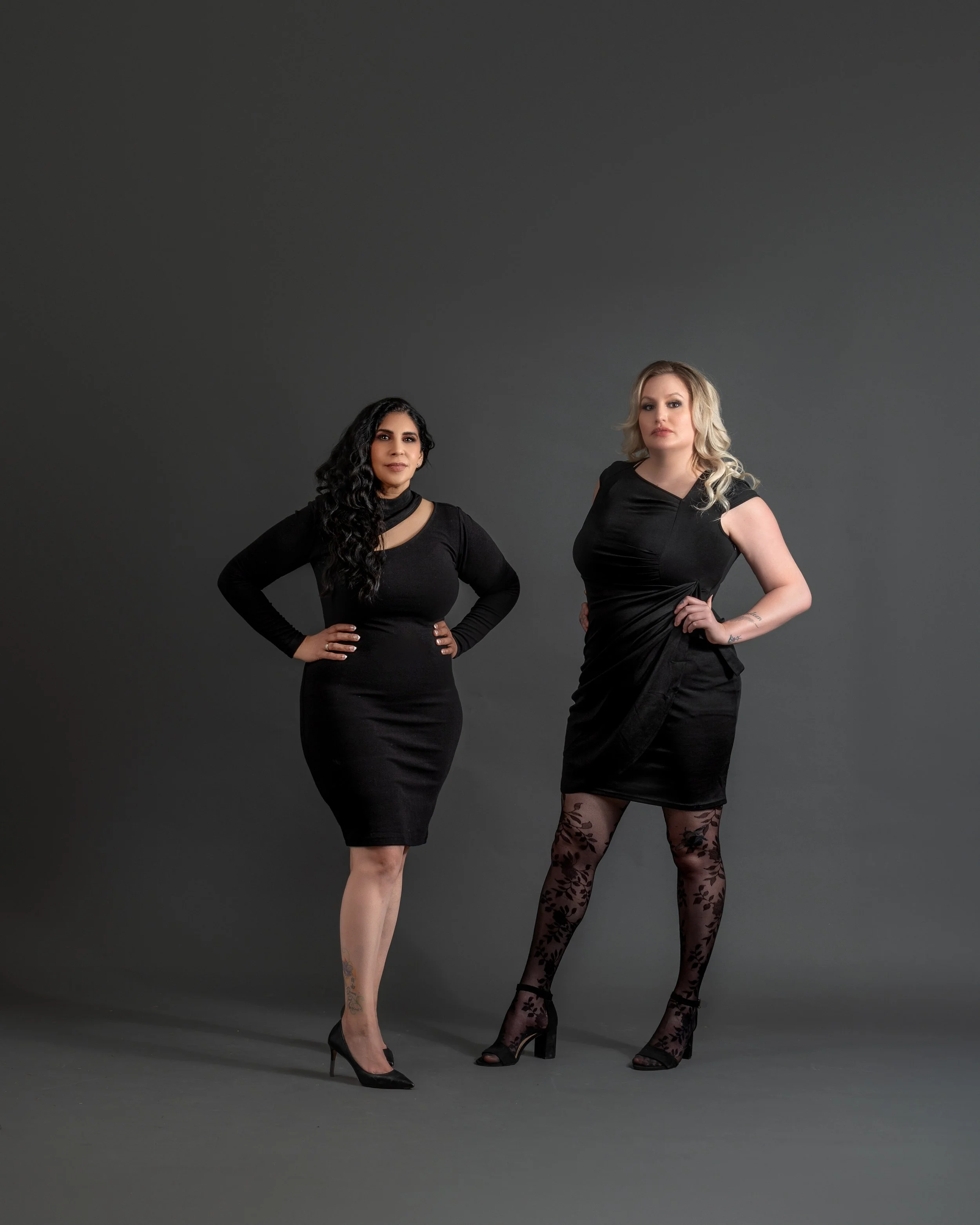 Two women standing side by side against a gray background, both wearing black dresses and high heels, with confident poses.