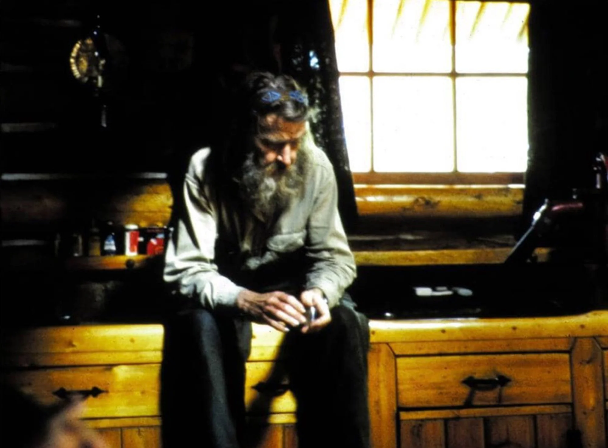 Wendell Beckwith with a long gray beard sits on a wooden counter top in the kitchen of one of his cabins
