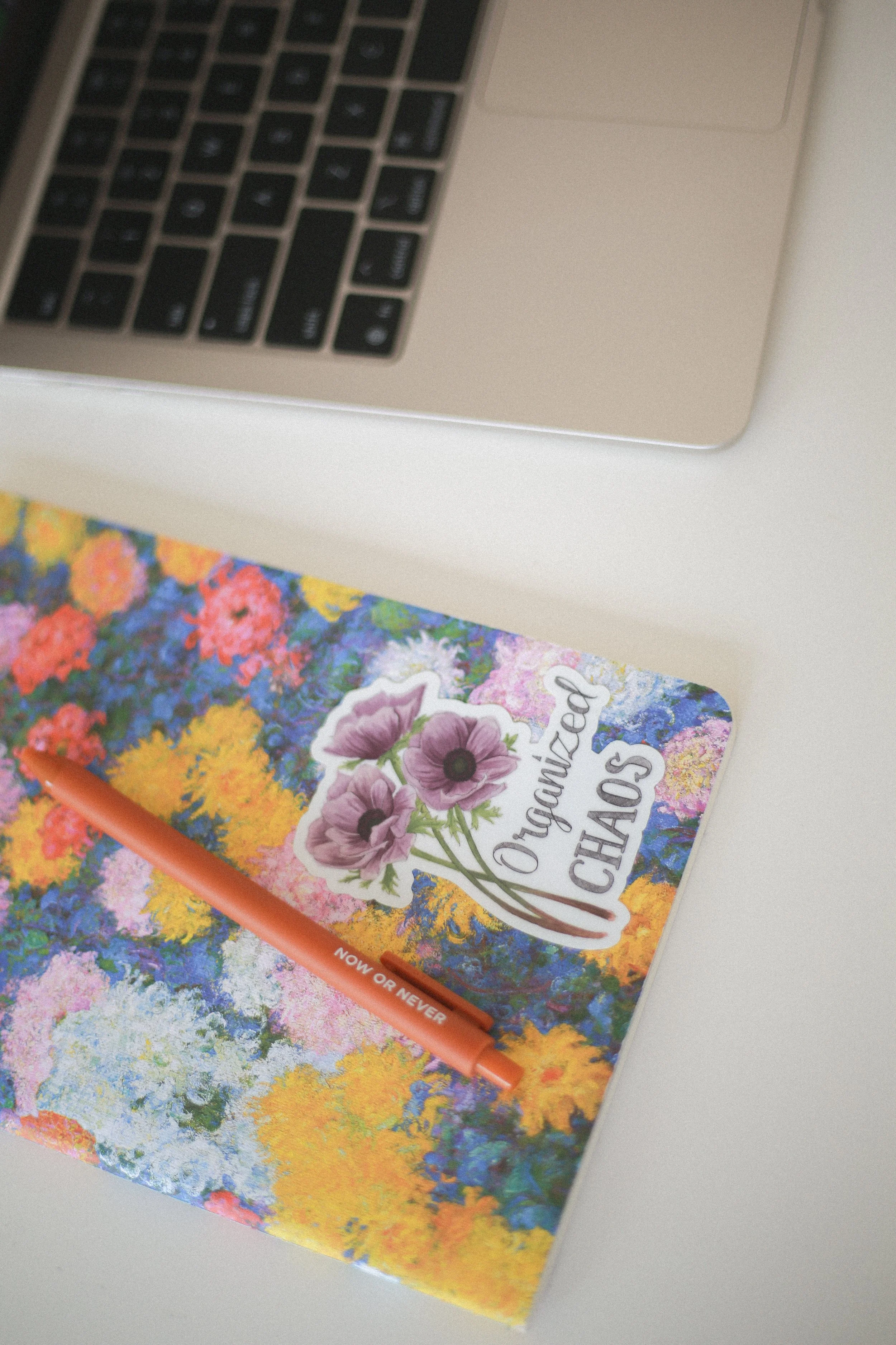 A colorful floral-patterned notebook with a sticker featuring purple flowers and the words 'Oregano Chips'. An orange pen lies on top of the notebook, and part of a laptop keyboard is visible in the background.