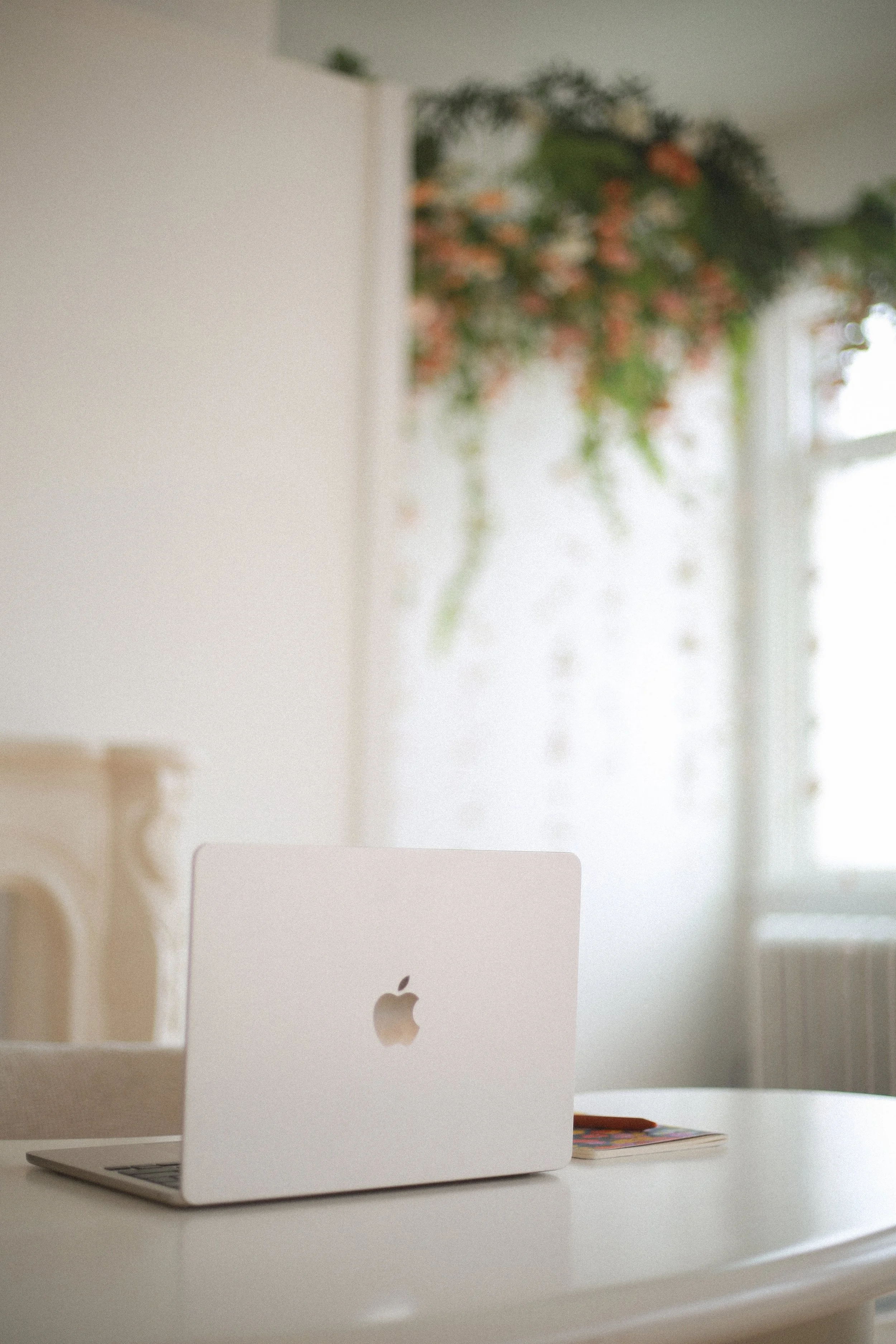 A white Apple iMac computer on a white desk with a colorful magazine underneath it. A window with a blurred view of greenery outside is in the background.