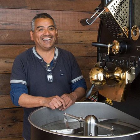 Man smiling while standing next to a coffee roasting machine in a rustic wooden setting.