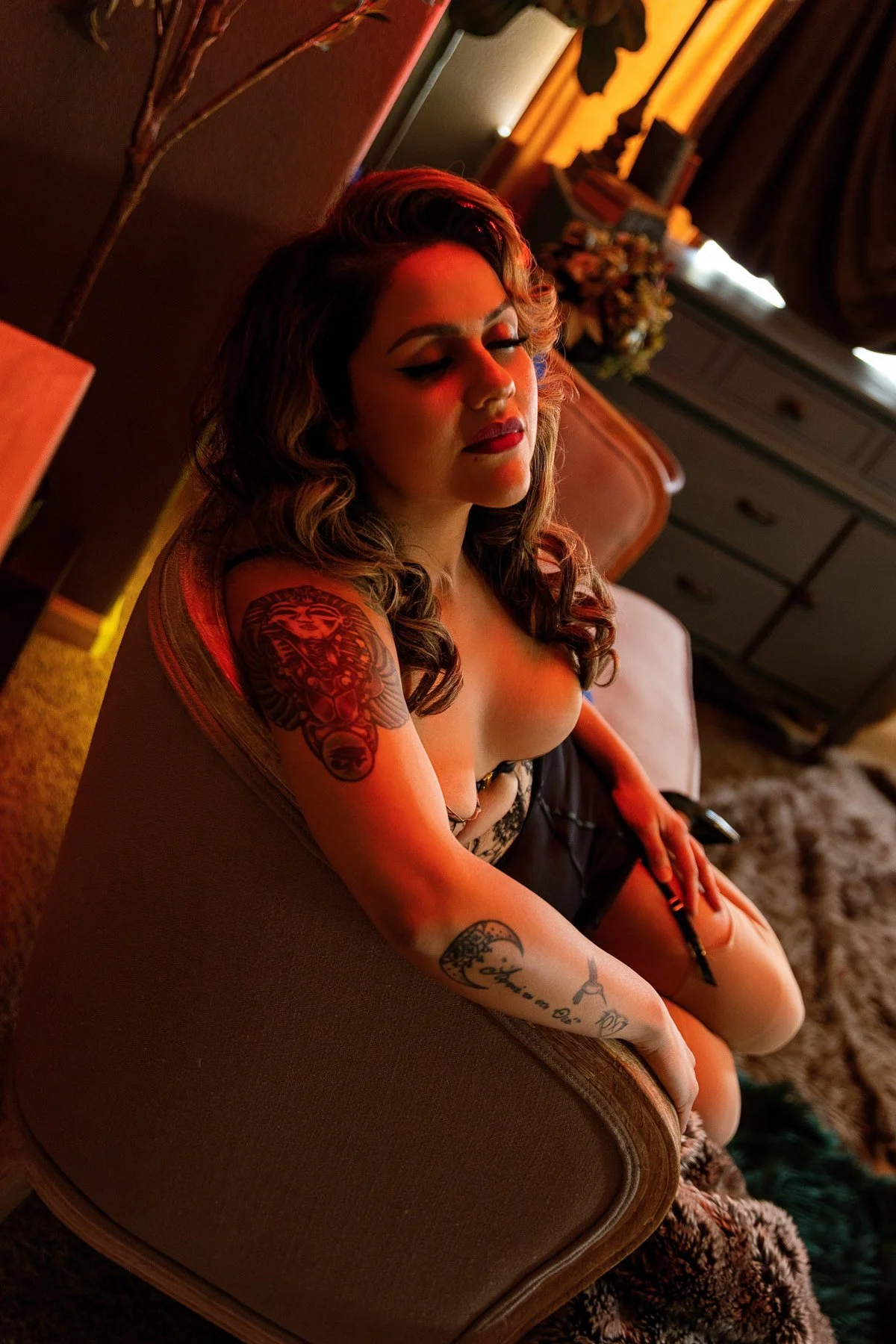 A young woman with styled hair and makeup, sitting on a brown upholstered chair with her eyes closed. She has tattoos on her arm and is wearing a revealing top, in a dimly lit room with warm lighting.