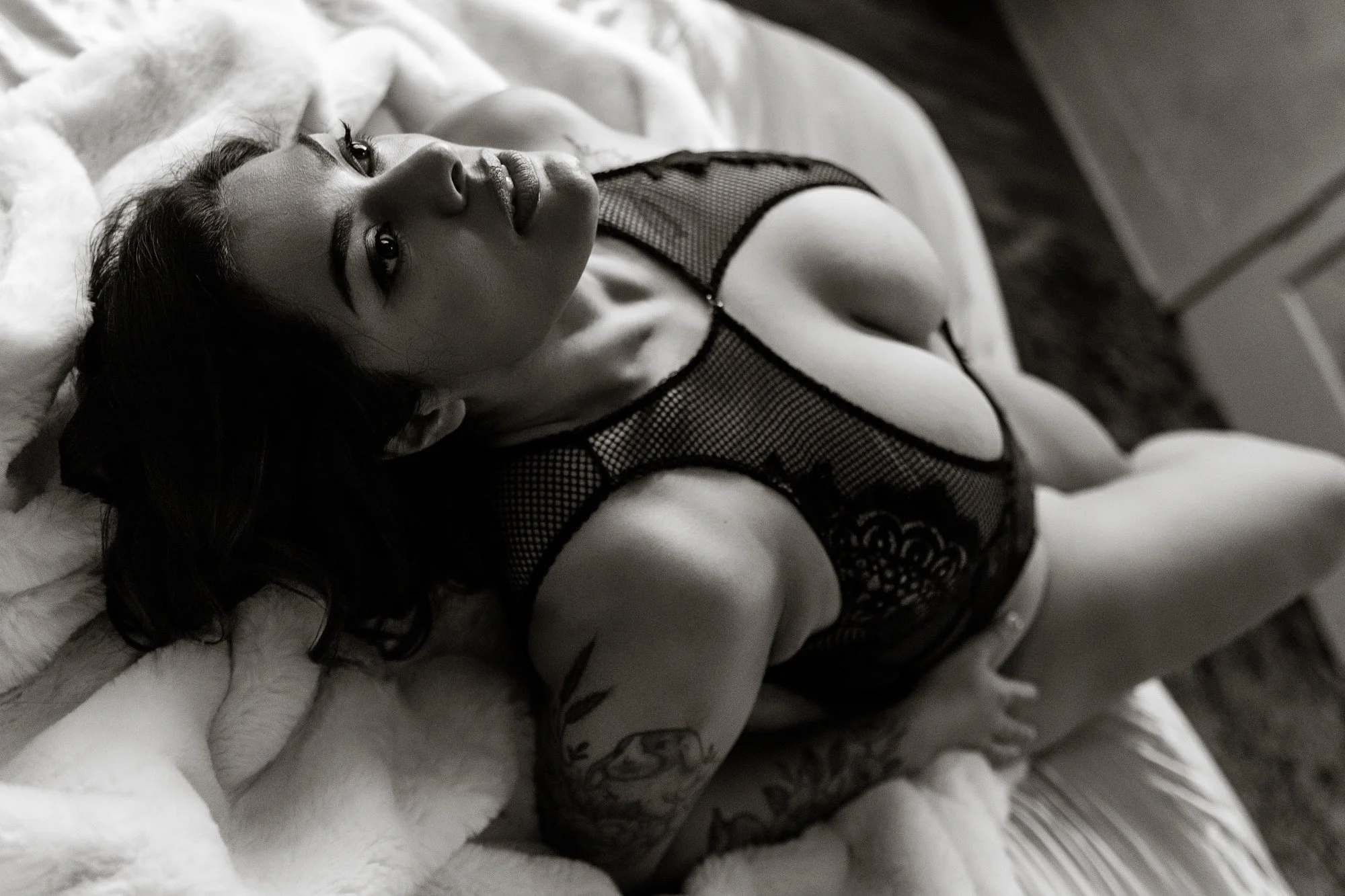Black and white photo of a woman lying on a bed, looking up at the camera. She is wearing a fishnet top and patterned shorts, with long dark hair spread out around her.