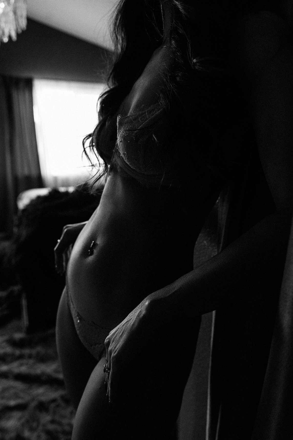 A black and white photo of a woman in lingerie, with focus on her stomach and side profile, in a dimly lit room with a window in the background.