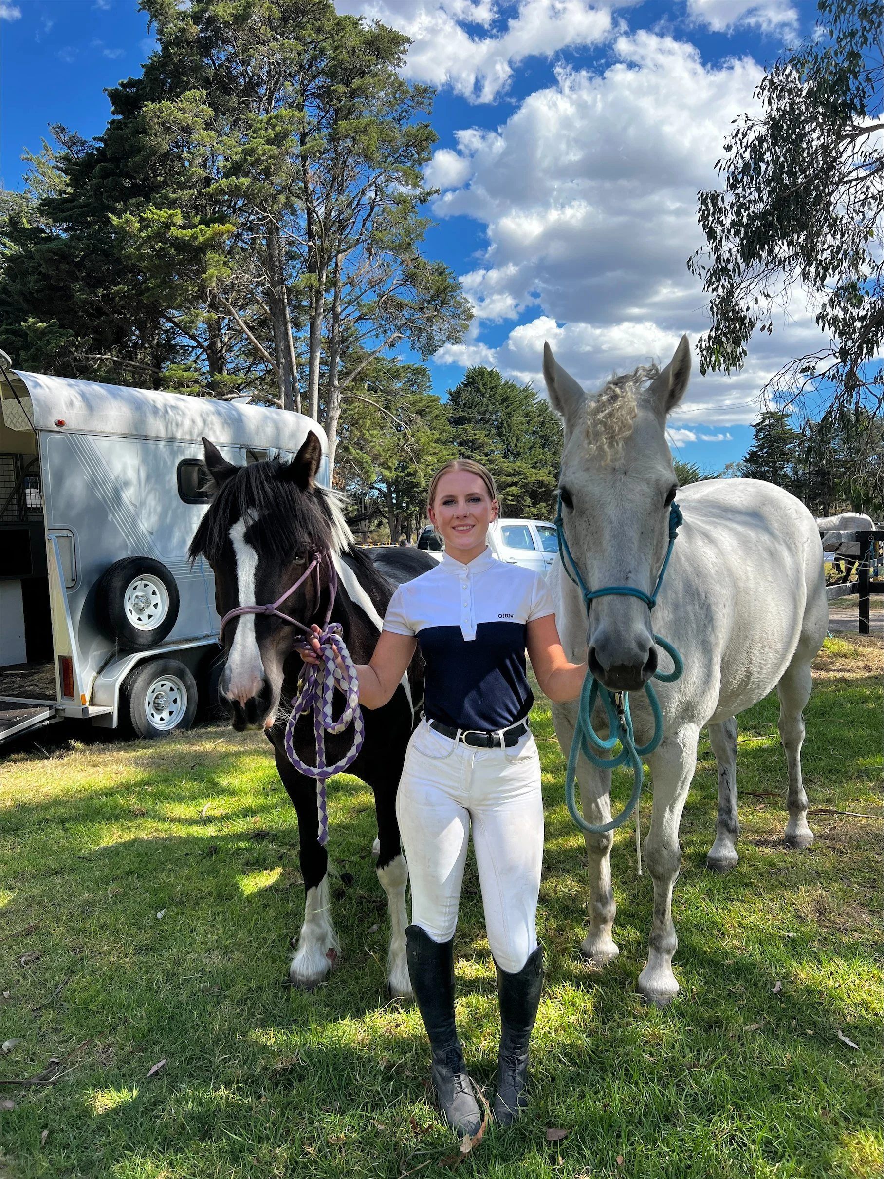One of our nurses standing outdoors on grass, holding two horses, one black and white and the other white, with a trailer and trees in the background on a partly cloudy day. Blackburn Animal Hospital