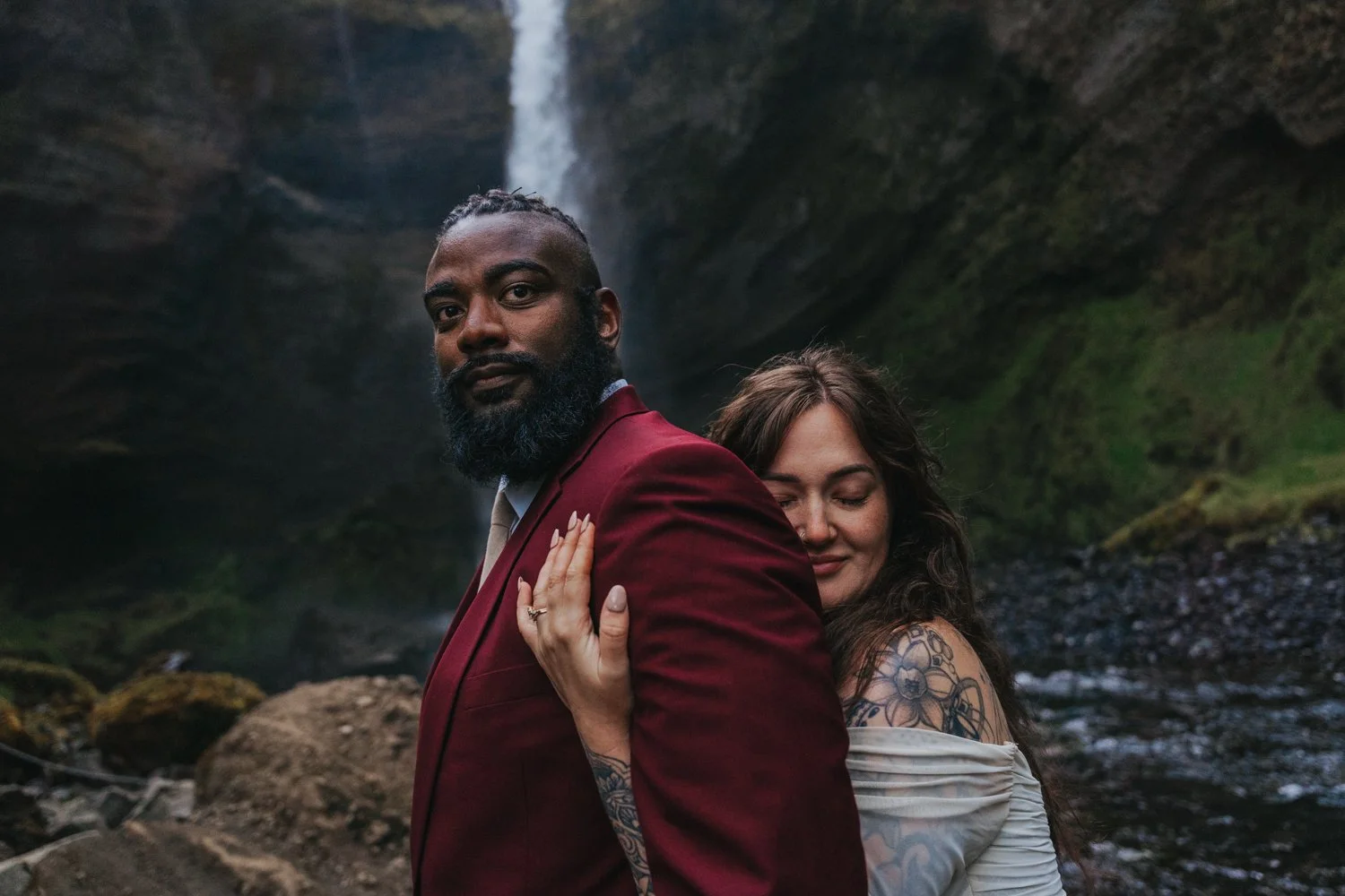 Couple eloping in Iceland. Bride hugs her groom from behind as they stand in front of a majestic waterfall