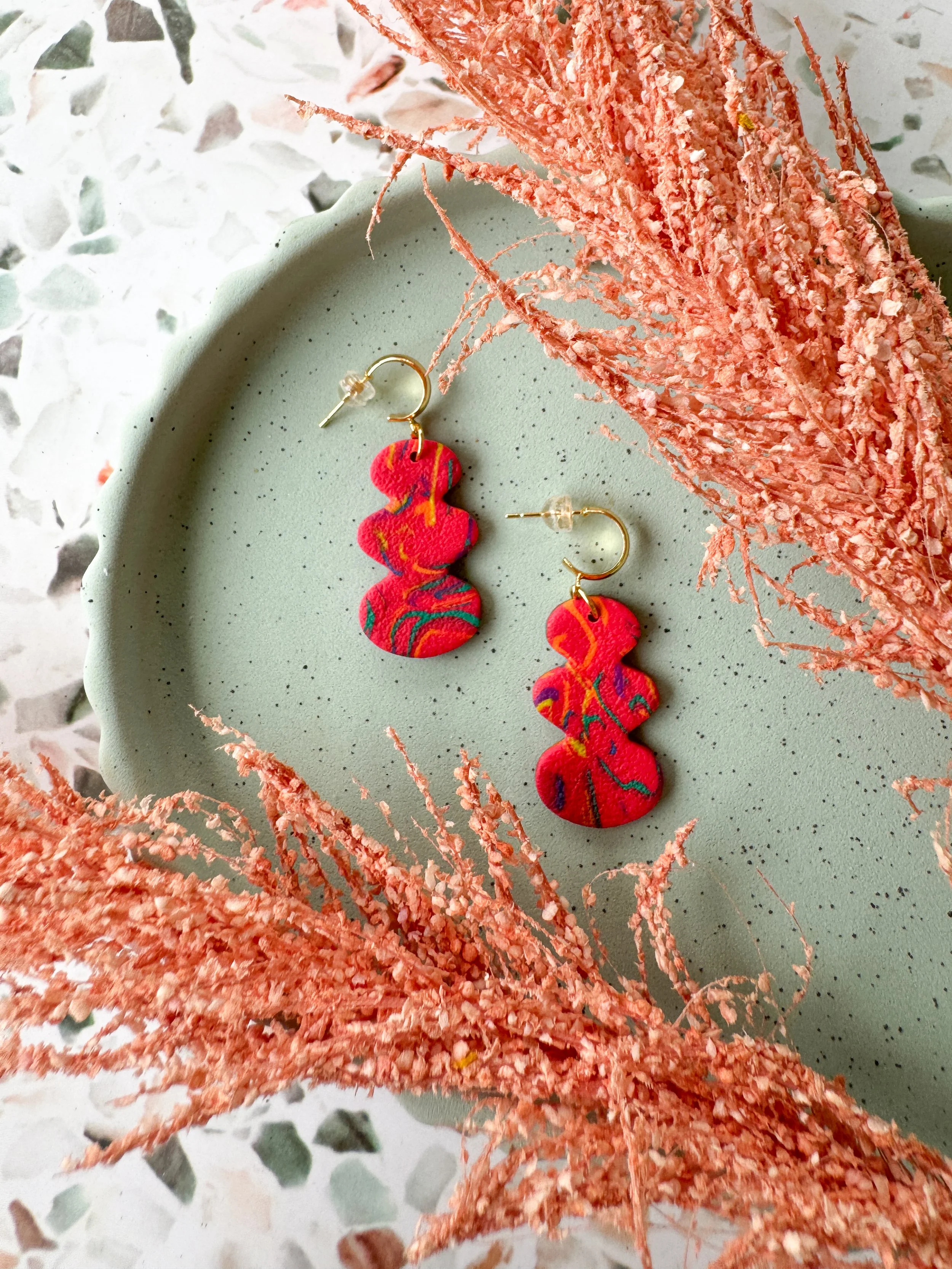 Pair of colorful abstract-patterned earrings on a green dish, surrounded by pink dried decorative foliage.