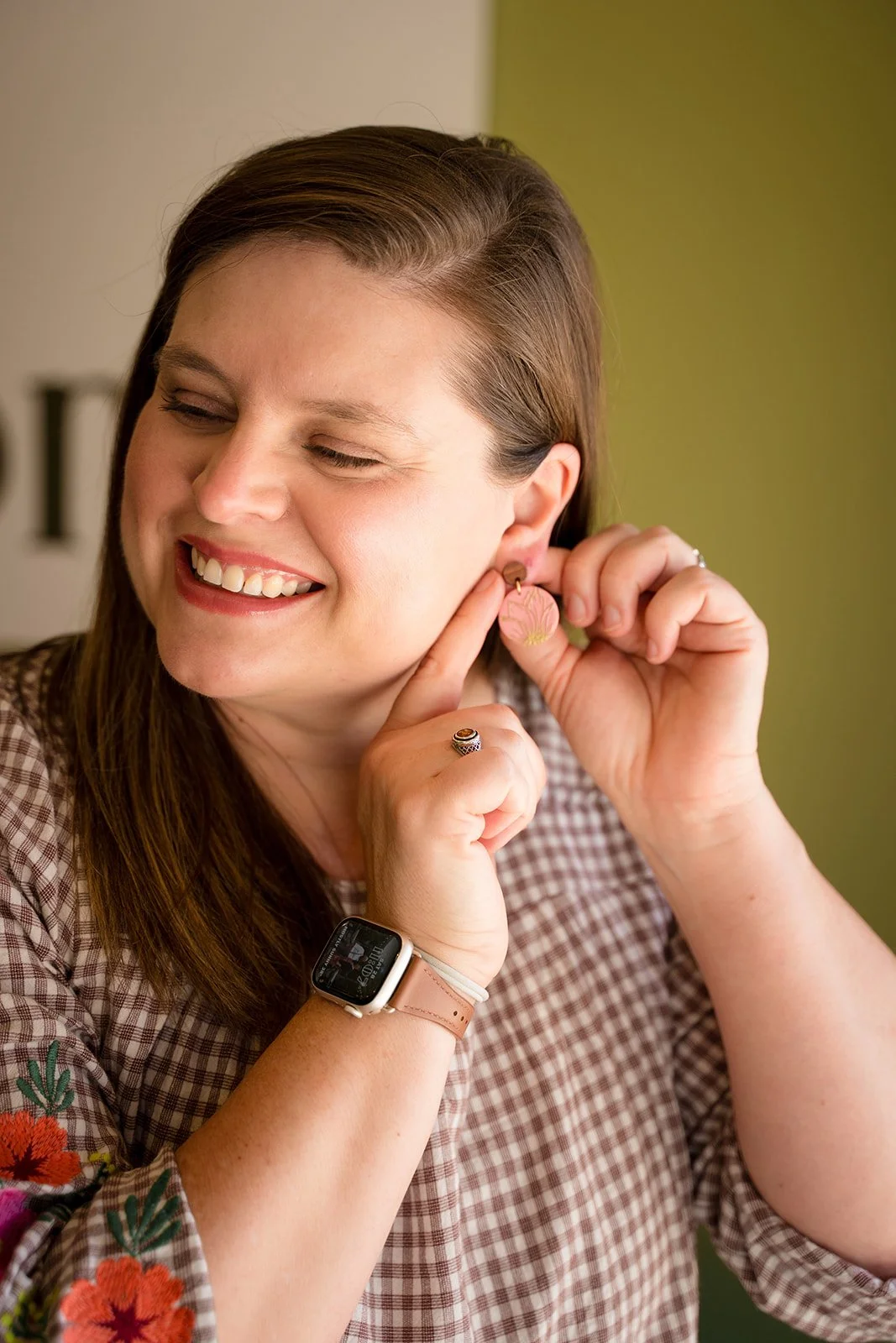 Julie Mullins, maker and owner of Anam Cara Clay Goods, smiling while putting on a pair of handmade polymer clay earrings against a sage green background.