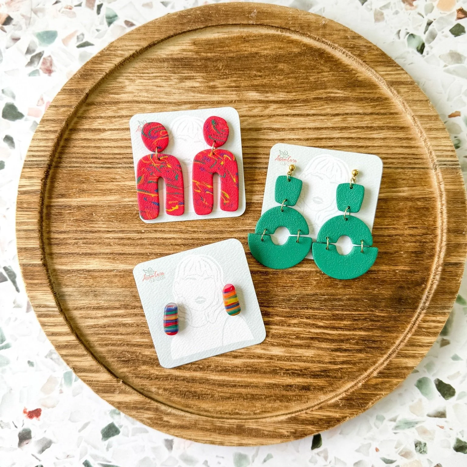 Three pairs of colorful earrings on white display cards placed on a round wooden tray.