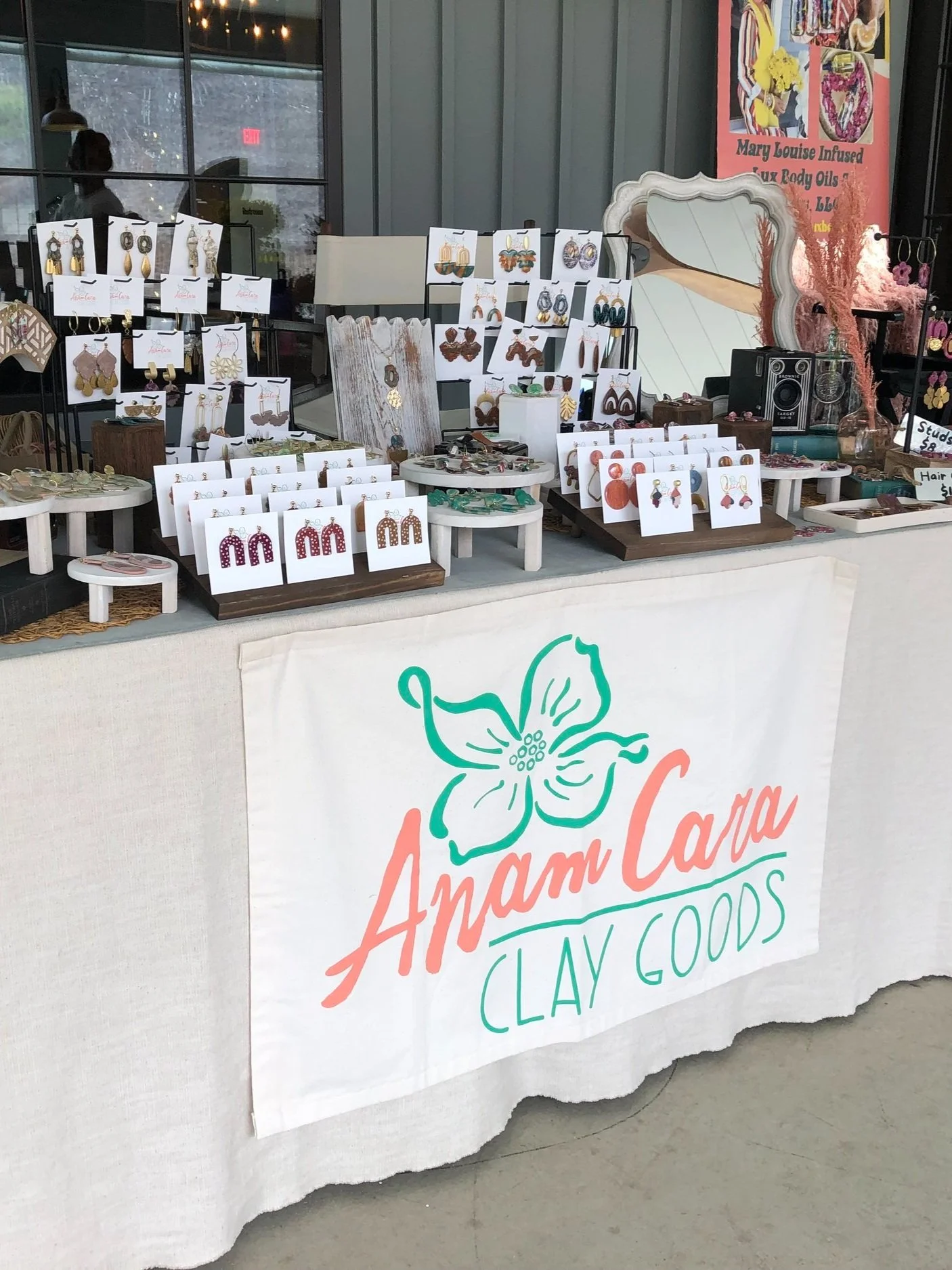 A market stall displays handmade earrings and jewelry, with a cloth banner reading "AmamCarla Clay Goods" and a flower illustration.