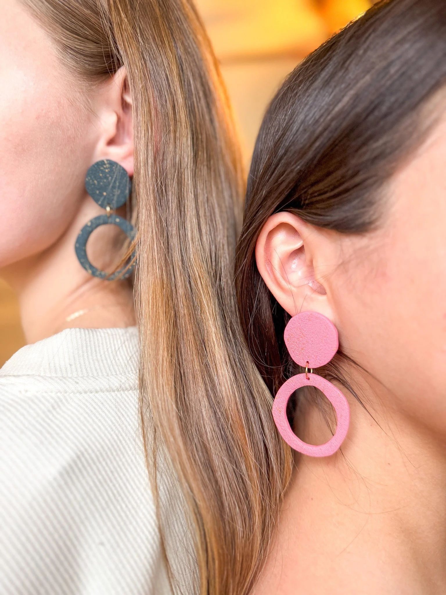 Close-up of two women wearing large, circular statement earrings, with one woman in blue earrings and the other in pink earrings.