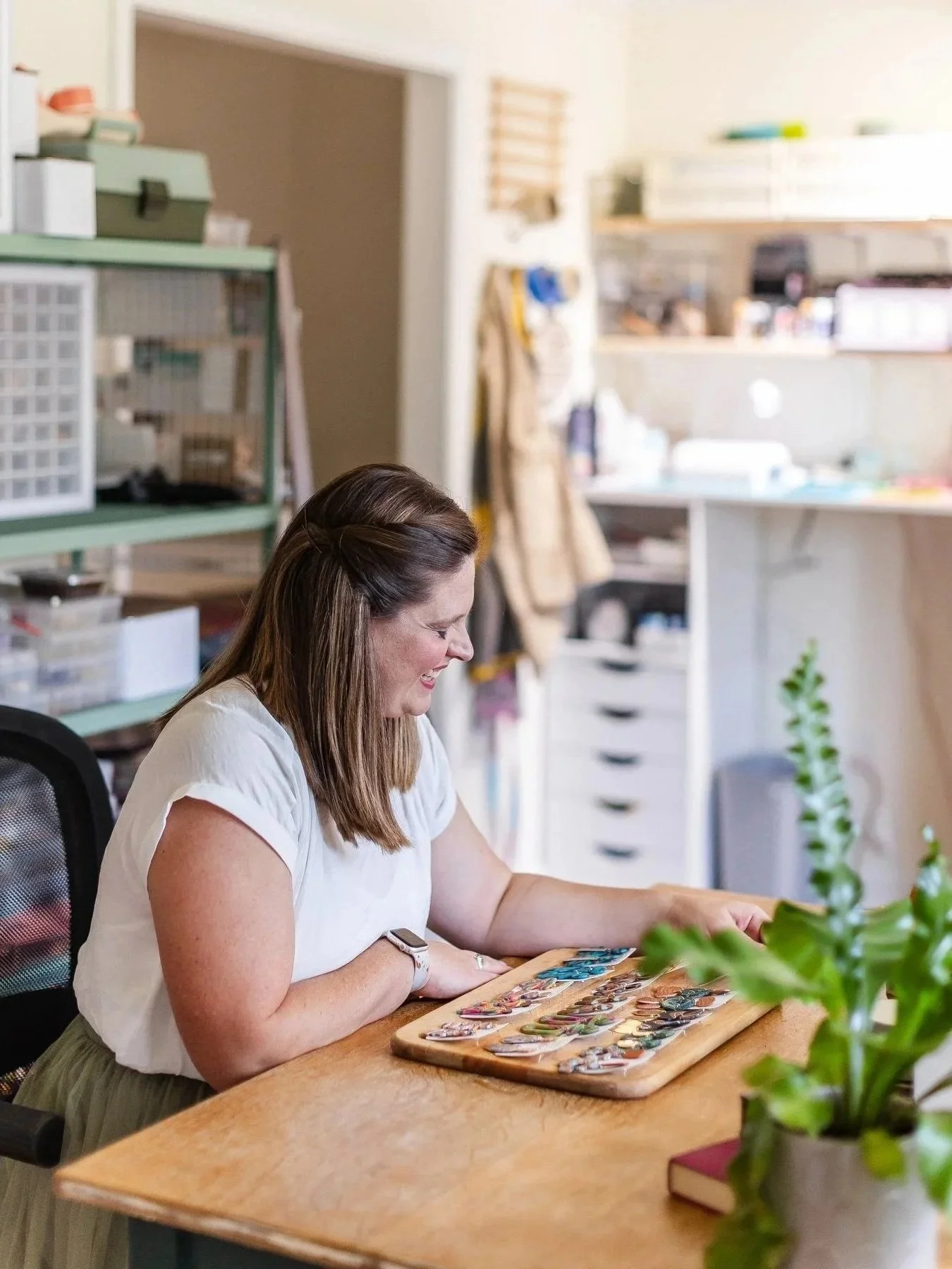 A woman with brown hair sitting at a wooden table, smiling and looking at a display of jewelry or accessories. The background shows a cluttered workspace with shelves and storage boxes.