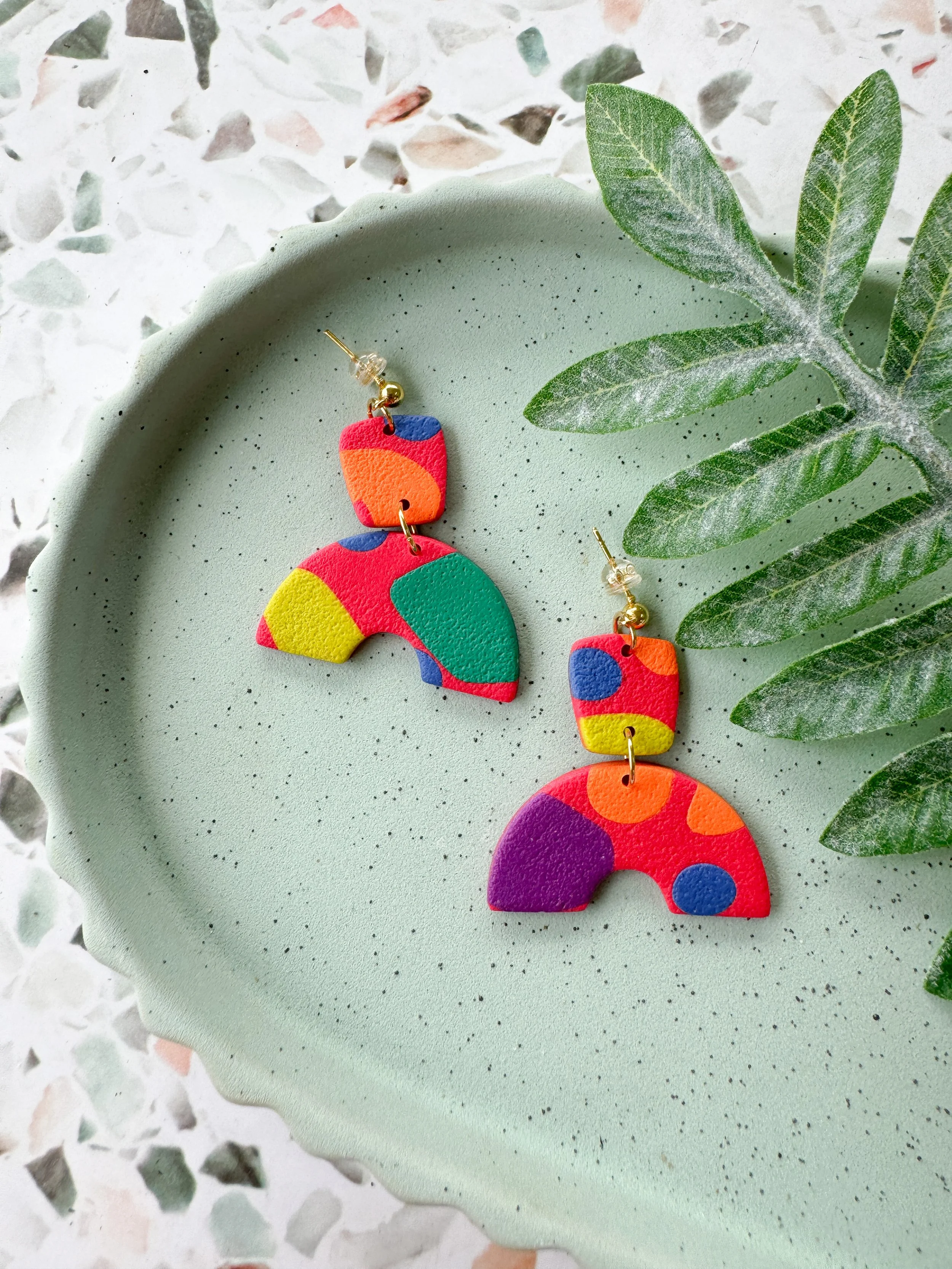 Colorful abstract earrings with geometric shapes on a speckled green plate, next to a green leaf.