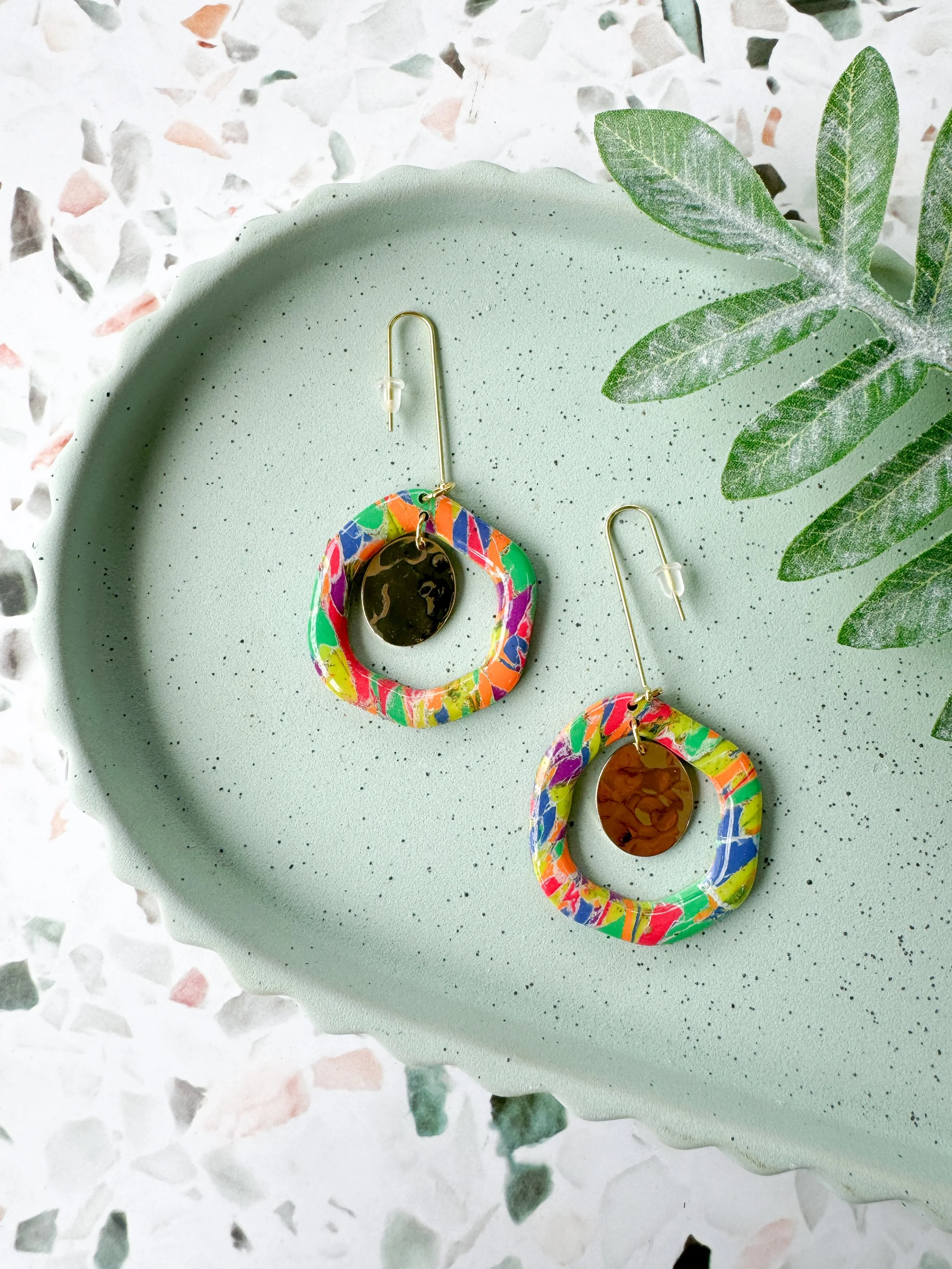Colorful earrings with circular hoops and dangling oval charms on a mint green dish, with green leaves nearby on a terrazzo surface.