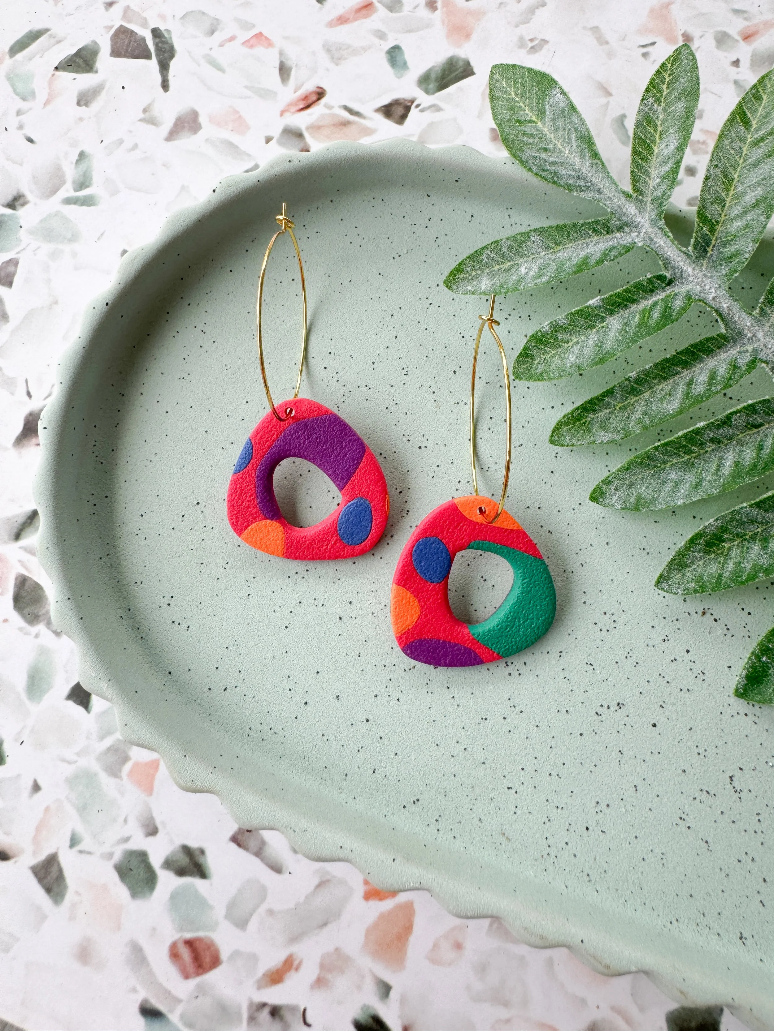 Colorful, abstract earrings with oval shapes and vibrant multicolor patterns, placed on a speckled green ceramic plate with a green leafy branch nearby.