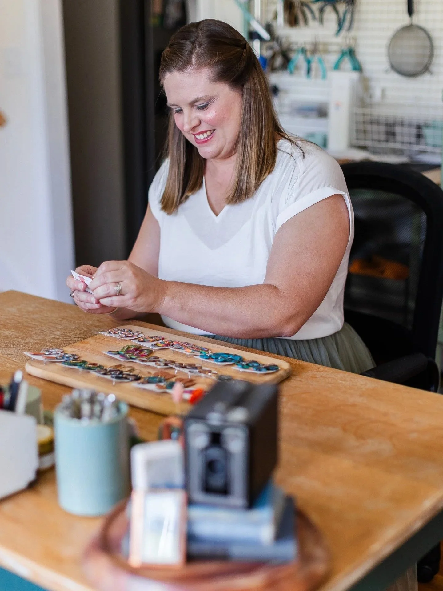 A woman with brown hair smiling while working on colorful jewelry pieces at a wooden table in a craft room.