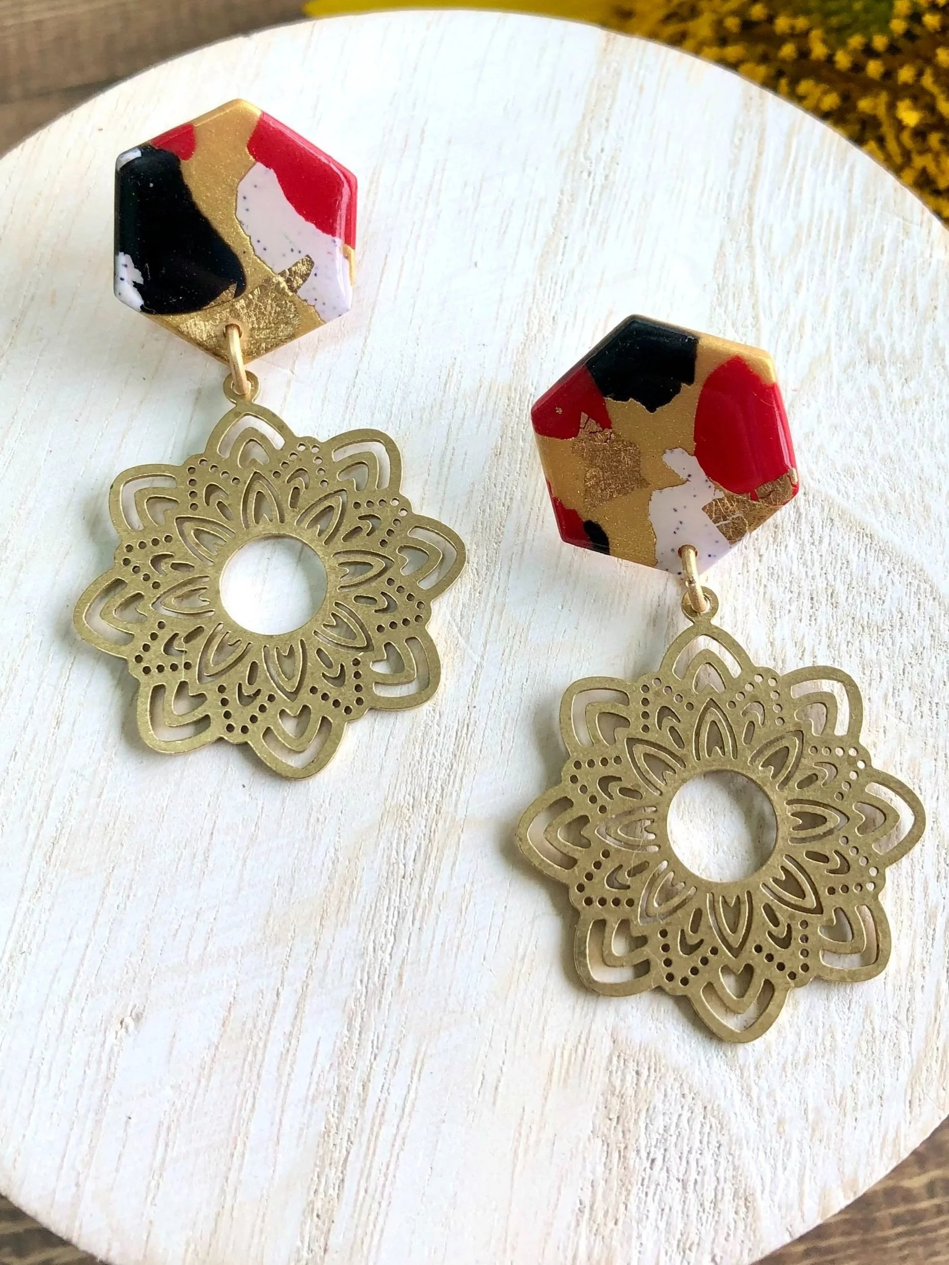 Pair of earrings with hexagonal red, black, white, and gold patterned tops and gold filigree floral pendants on a white wooden surface.