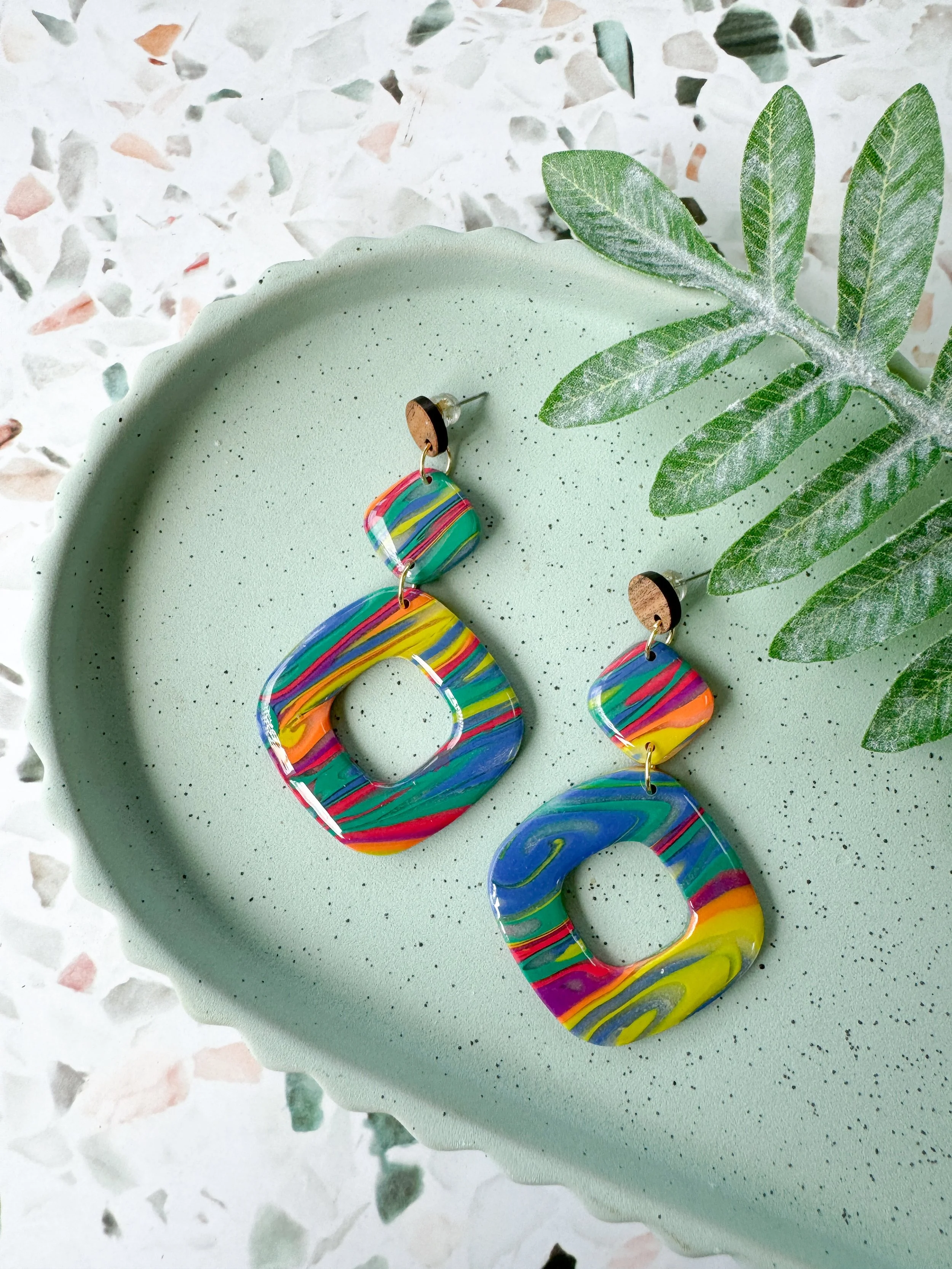 Colorful polymer clay earrings with a marbled rainbow pattern on a light green speckled dish, with green fern leaves nearby.