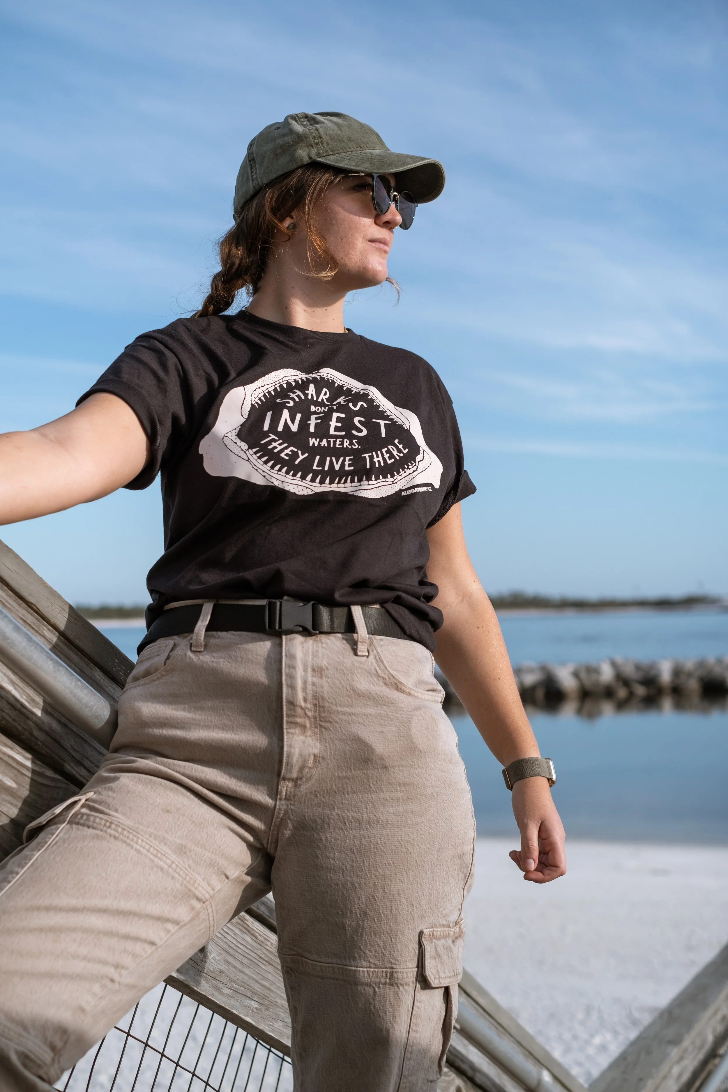 A person wearing a black t-shirt with a shark graphic and text, khaki cargo pants, a green cap, sunglasses, and a watch, standing outdoors by a body of water with a clear sky in the background.